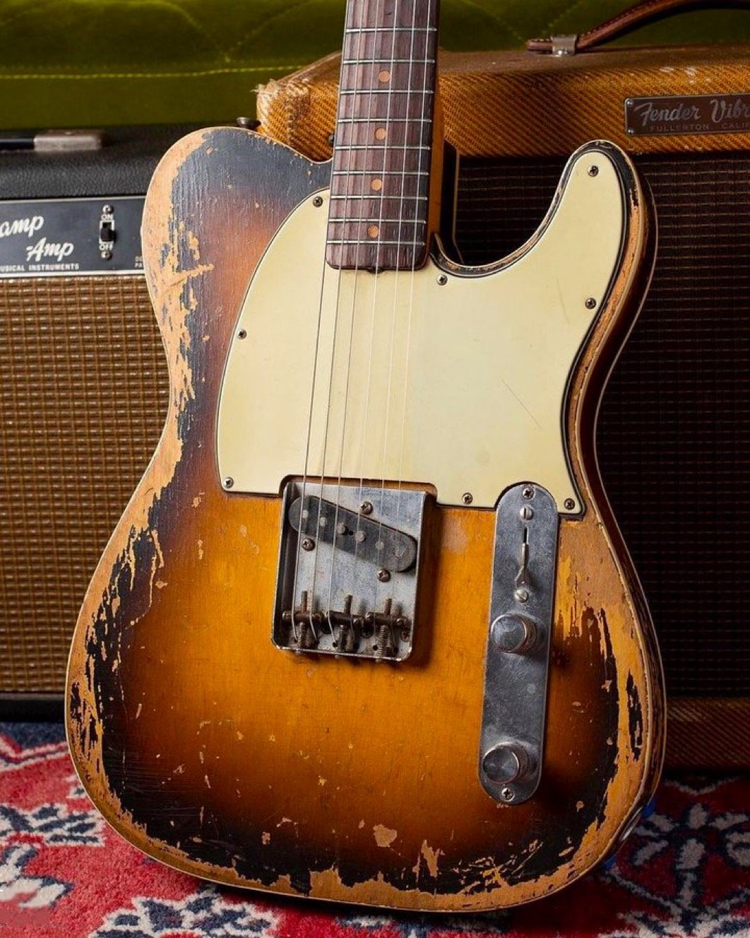Vintage electric guitar in front of a Fender amplifier on a patterned rug.