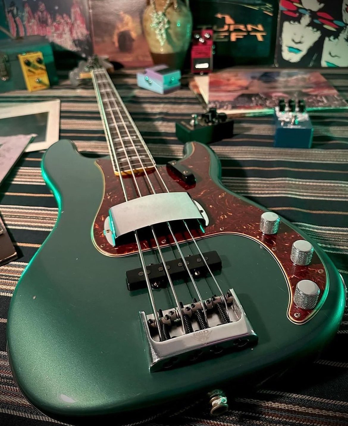 Close-up of a green electric bass guitar on a striped surface, surrounded by various music equipment and vinyl records in the background.