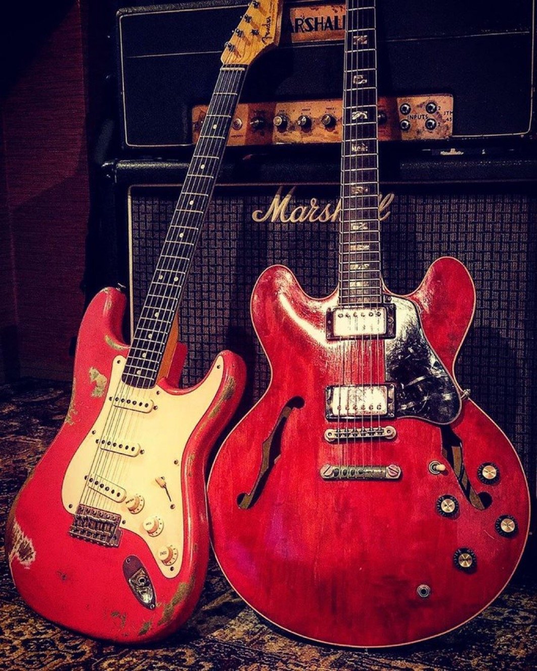 Two electric guitars, one Stratocaster-style and one semi-hollow body, both red, leaning against a black Marshall amplifier.