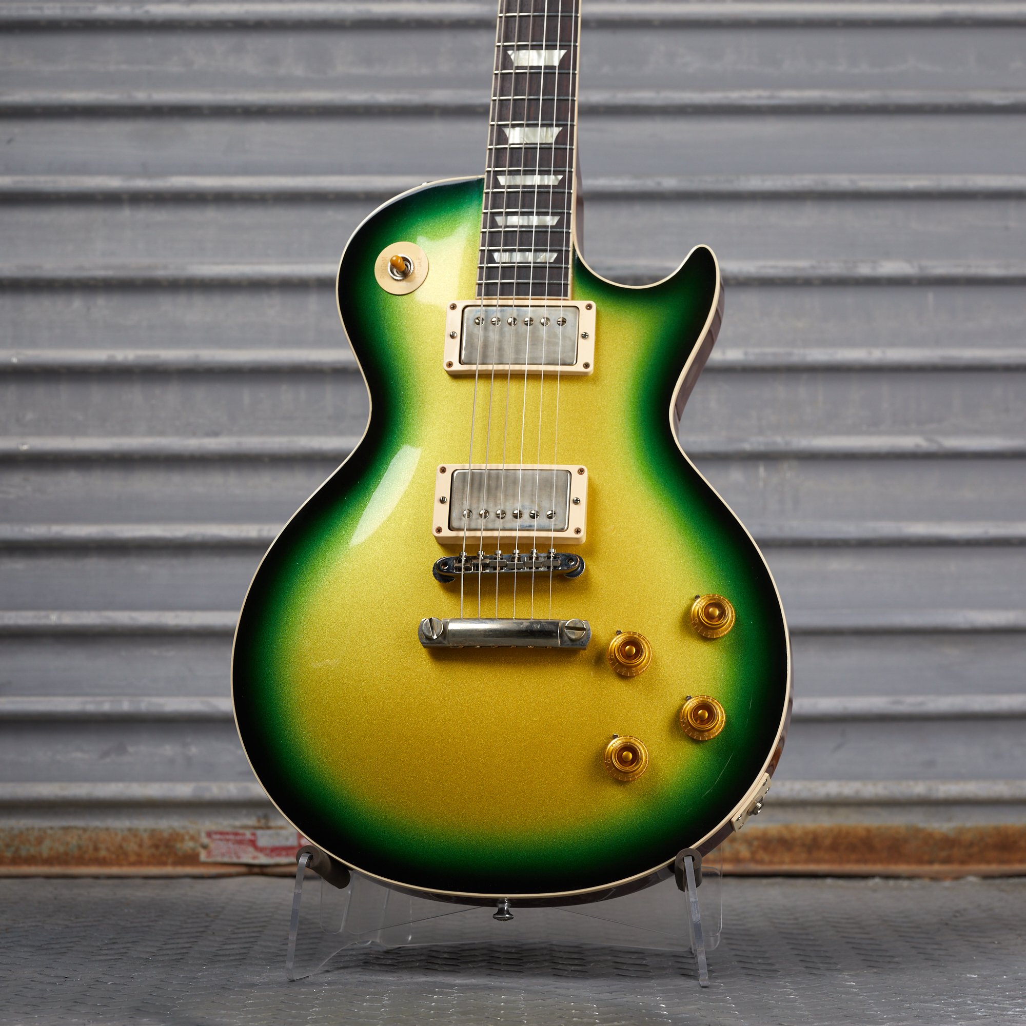 Electric guitar with green and gold gradient finish, silver pickups, and gold knobs, displayed against a corrugated metal background.