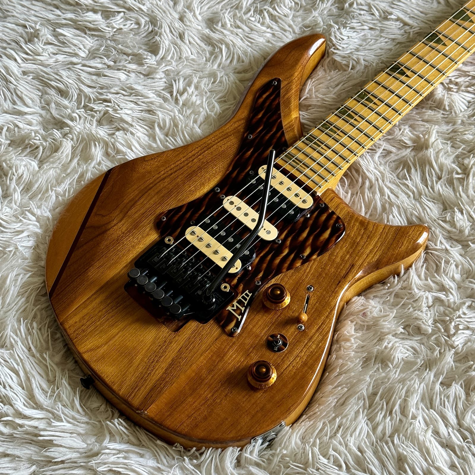 A wooden electric guitar with a carved body and intricate pickguard, lying on a textured white surface.