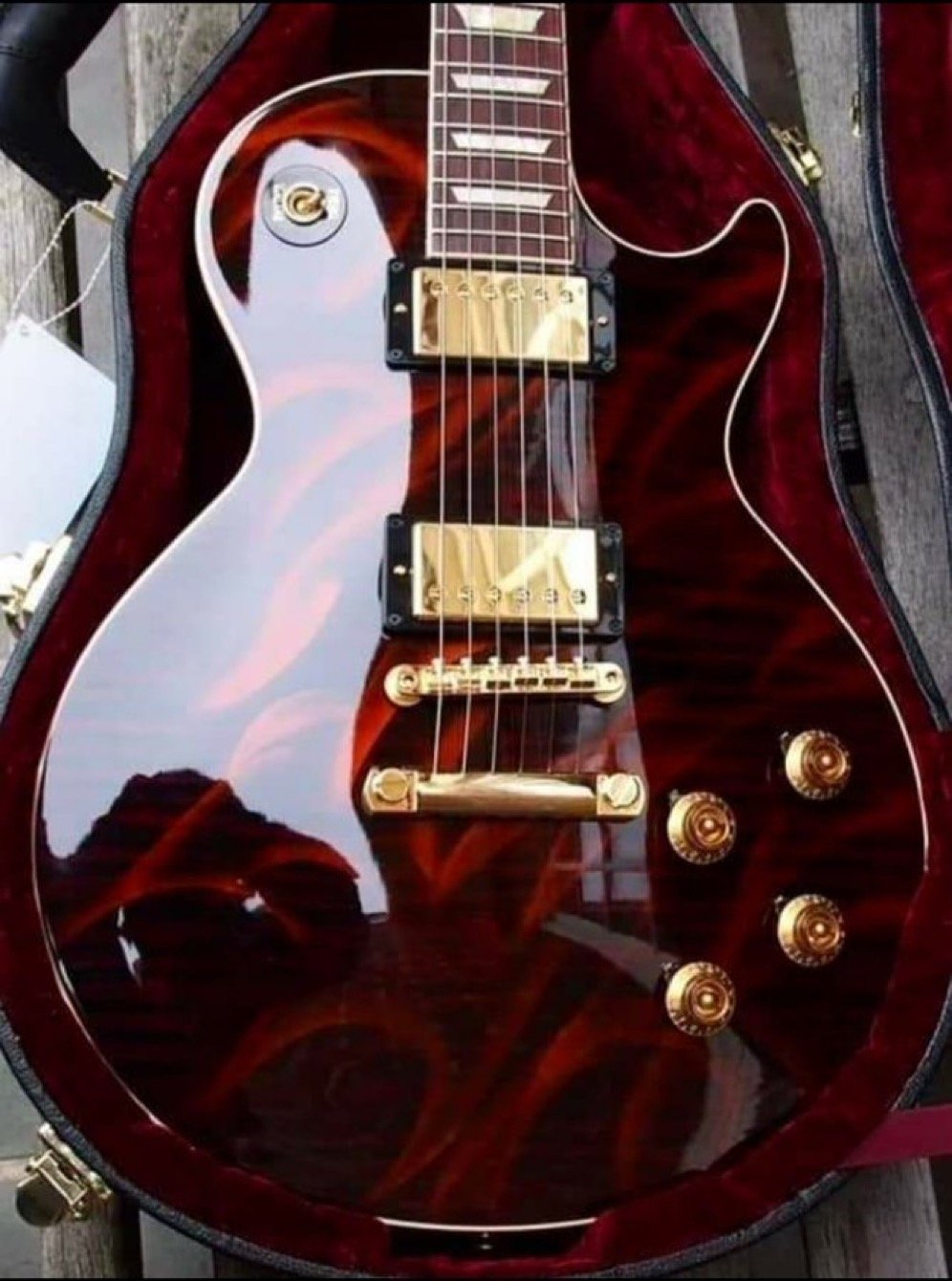 Electric guitar with a glossy flame maple finish and gold hardware, displayed in an open case. The guitar features two humbucker pickups and control knobs.