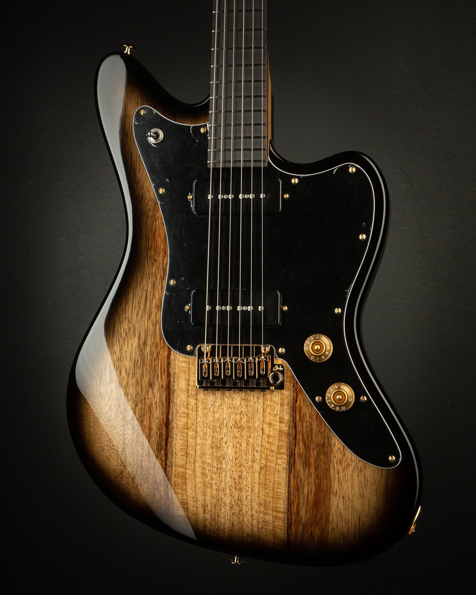 Close-up of an electric guitar with a brown wood finish and black pickguard on a dark background.