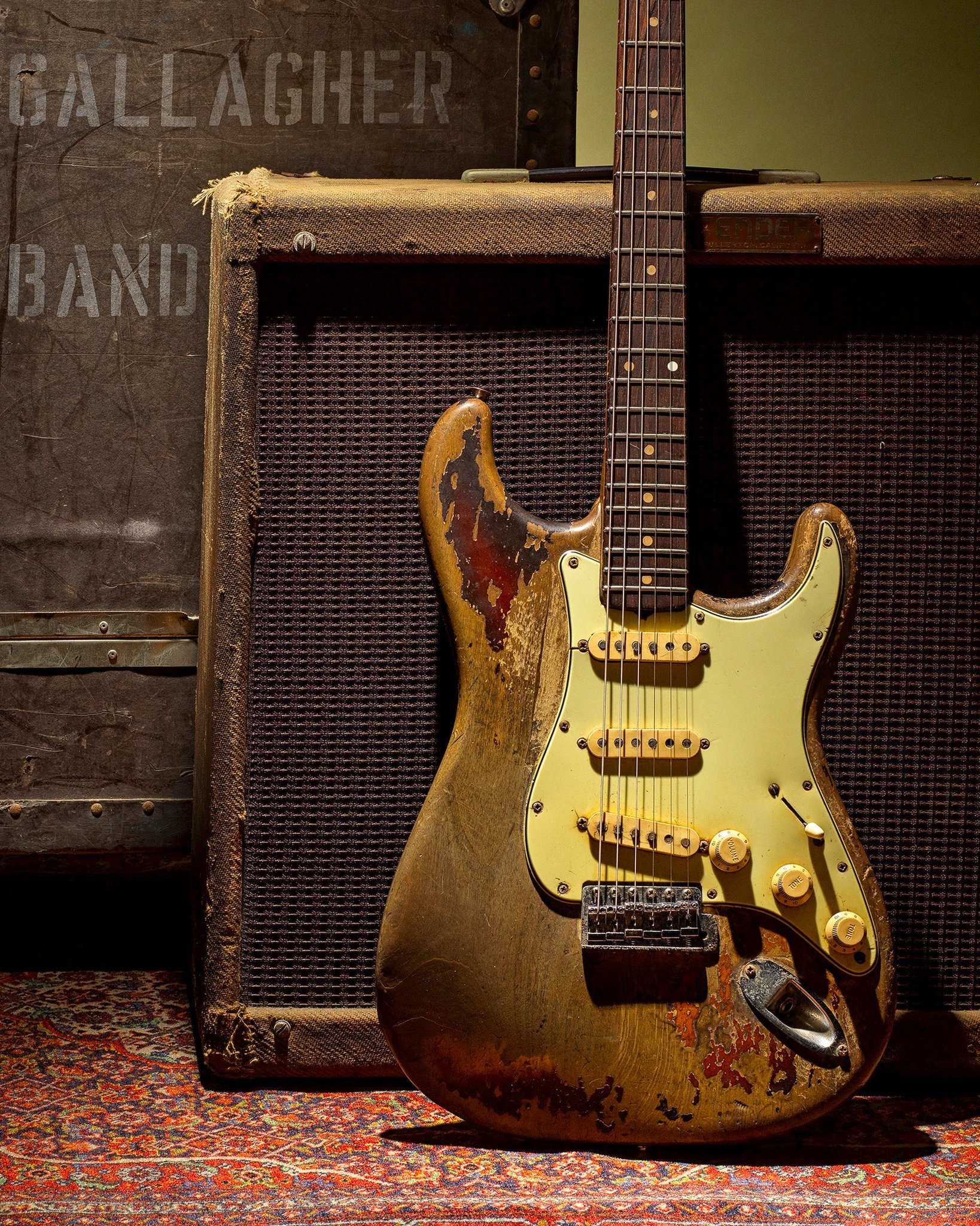 Worn electric guitar leaning against a vintage amplifier on a patterned rug.