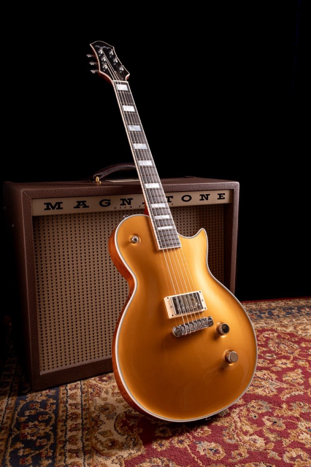 Electric guitar with gold finish leaning against a Magnatone amplifier on a patterned rug.