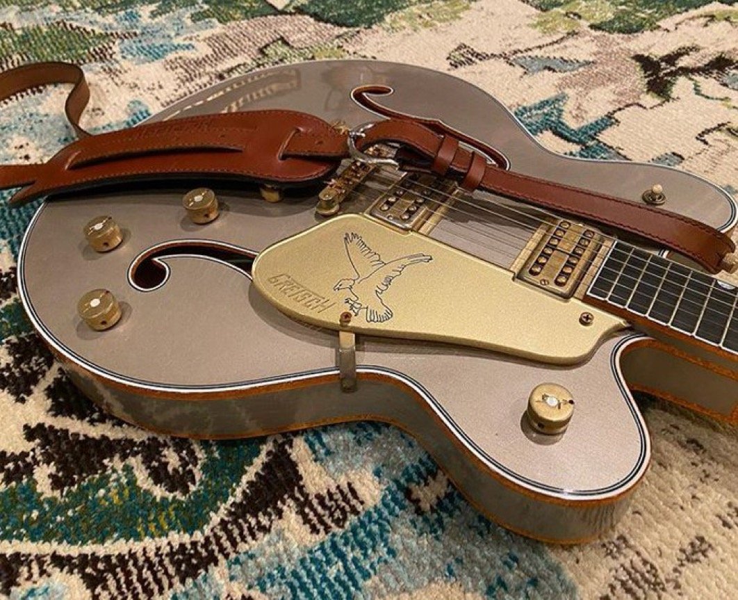 Silver Gretsch guitar with a brown leather strap, resting on a patterned rug. The guitar features a golden scratchplate with an engraved bird design.