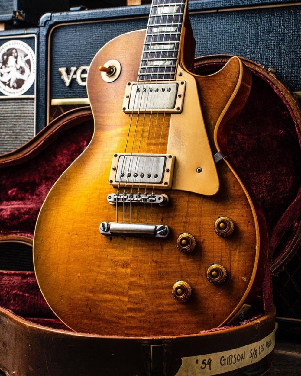 1959 Gibson Les Paul electric guitar with a sunburst finish in an open guitar case, surrounded by amplifiers.