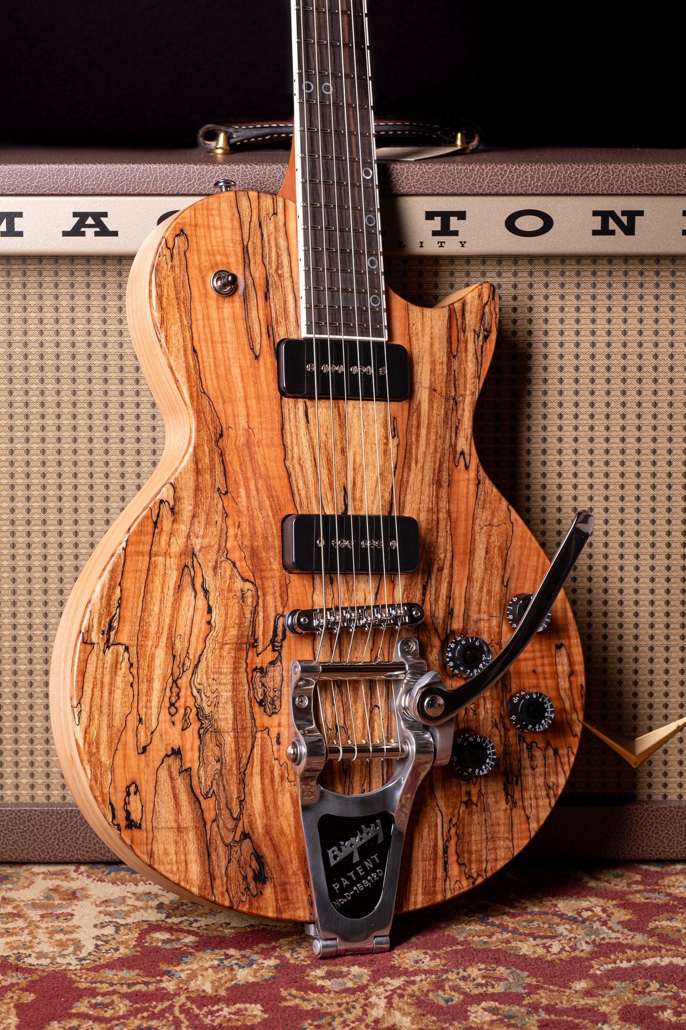 Electric guitar with spalted maple top, featuring a Bigsby tremolo, resting against an amplifier on a patterned rug.