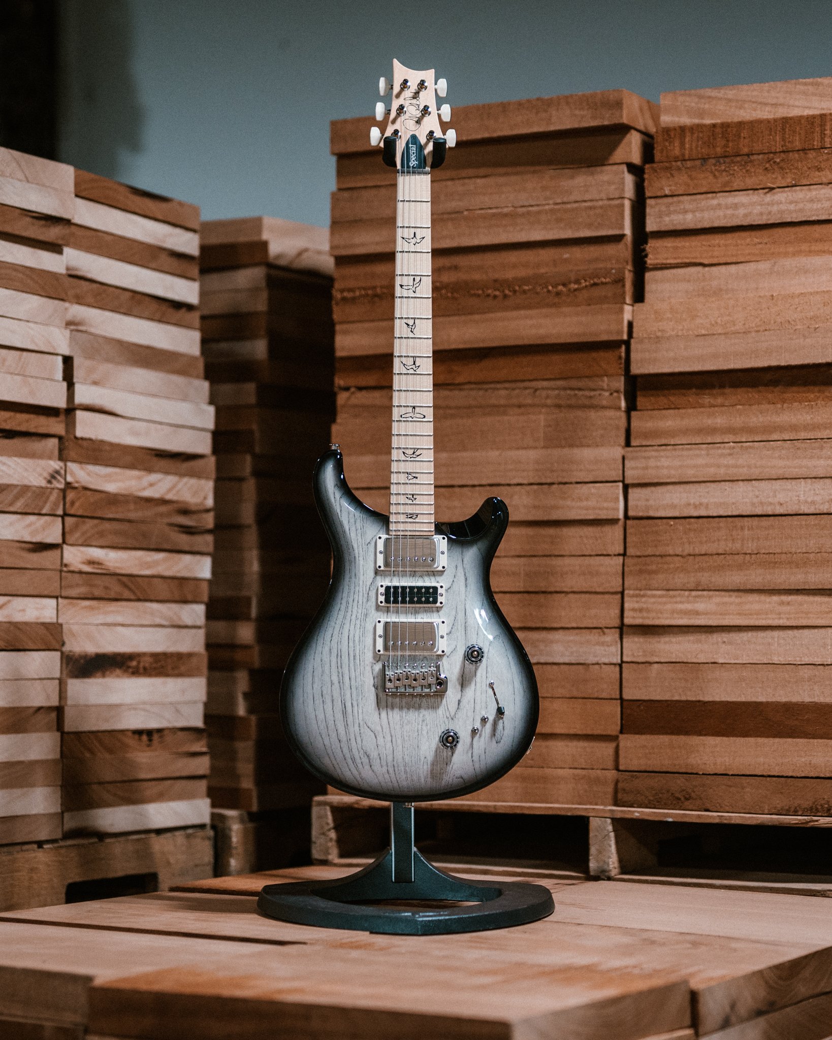 Electric guitar with bird inlays on fretboard, displayed on a stand, surrounded by stacked wooden planks.
