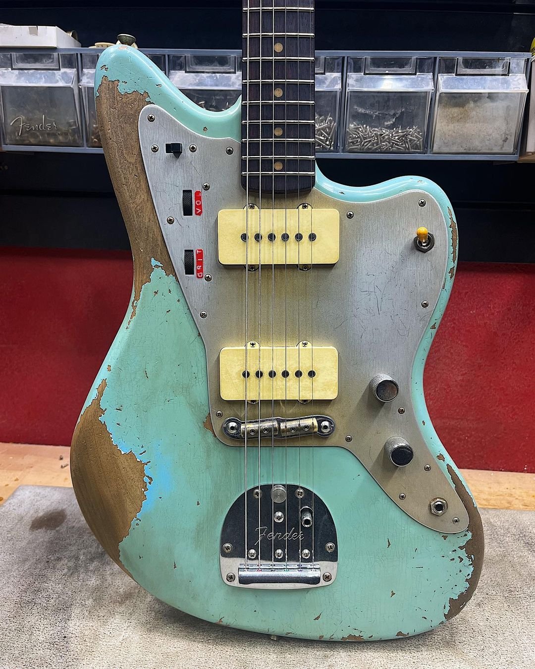 Close-up of a vintage electric guitar with a worn turquoise and wood finish, featuring two cream-colored pickups, a metal pickguard, and various control knobs and toggle switches. The background includes plastic storage containers with visible screws