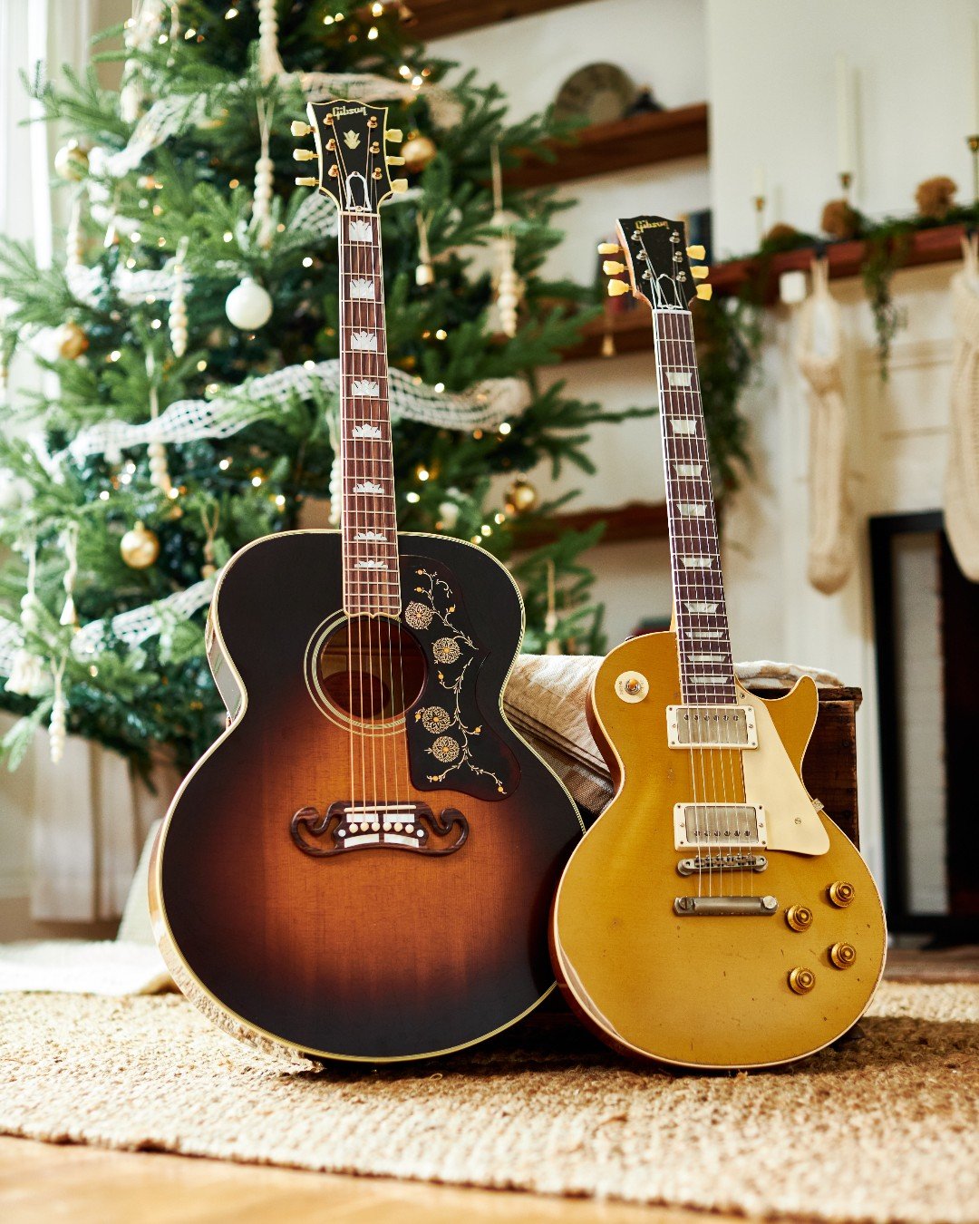 Two Gibson guitars in front of a decorated Christmas tree. One is an acoustic guitar with floral details, and the other is a gold electric guitar.