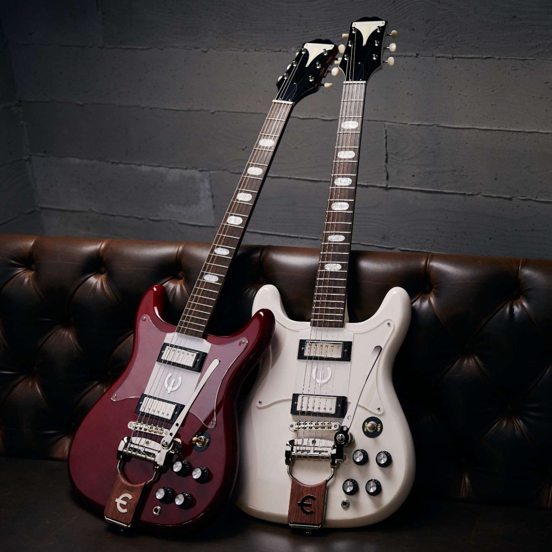 Two Epiphone electric guitars, one red and one white, leaning against a brown leather couch.