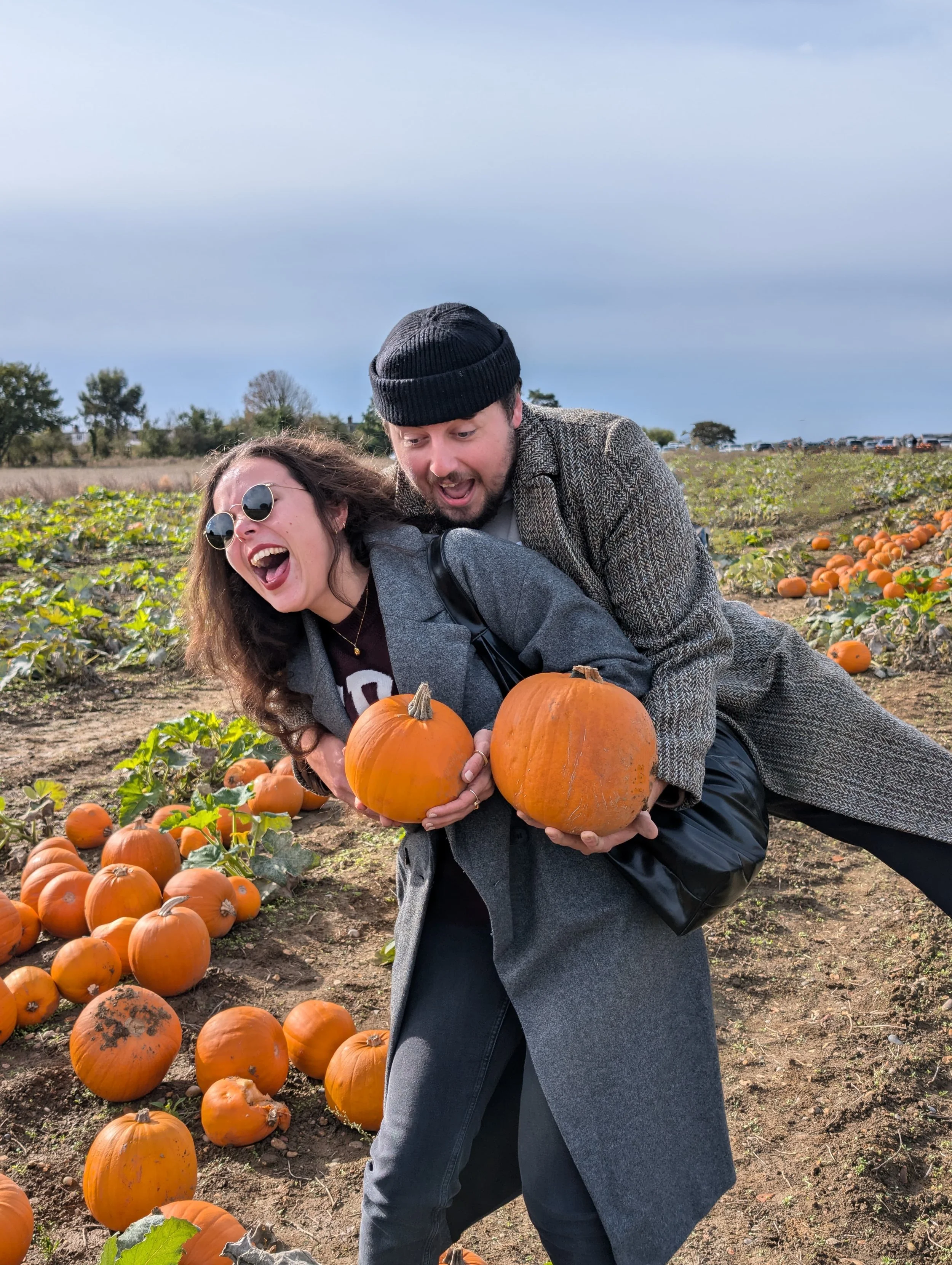 Two people, a man and a woman, smiling and holding pumpkins in a pumpkin patch during daytime.