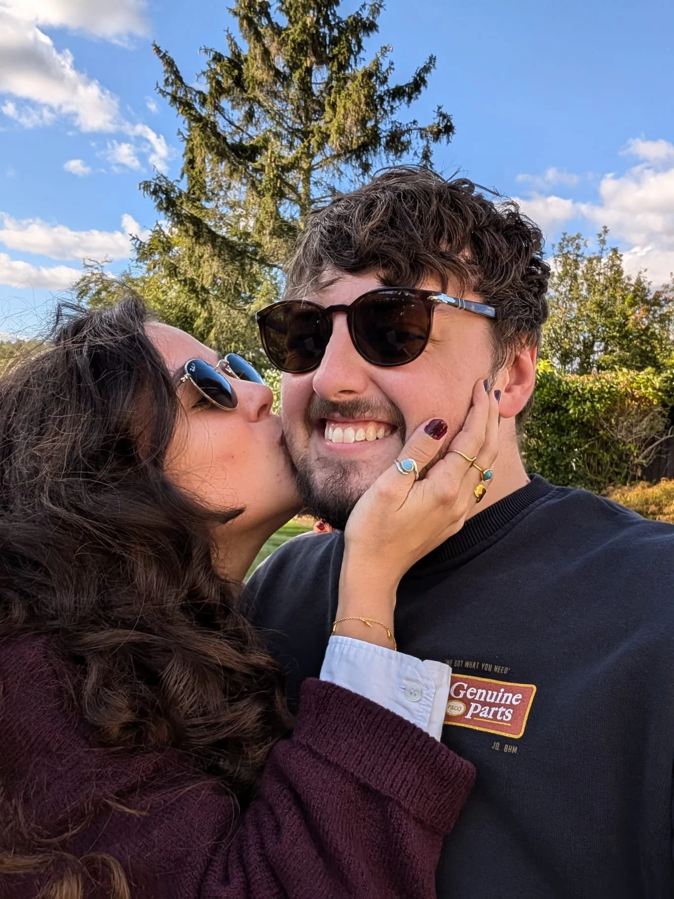 A woman with curly dark hair and sunglasses kissing a man with wavy dark hair and sunglasses on his face. The man is smiling and wearing a dark t-shirt with a logo that says "Genuine Parts." The woman has rings on her fingers and is holding the man's face. They are outdoors, with trees and a blue sky in the background.