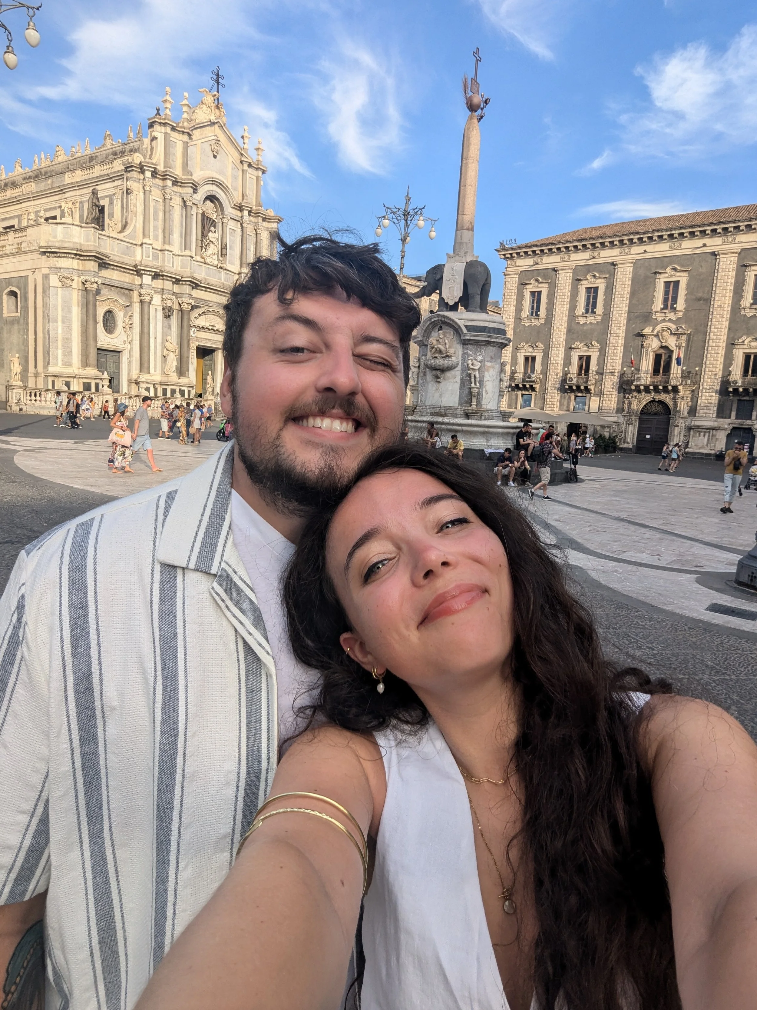 A smiling man with curly hair and a woman with long dark hair, both taking a selfie in a historic European square with a baroque church, an ancient Egyptian obelisk, and a fountain in the background, during daytime.