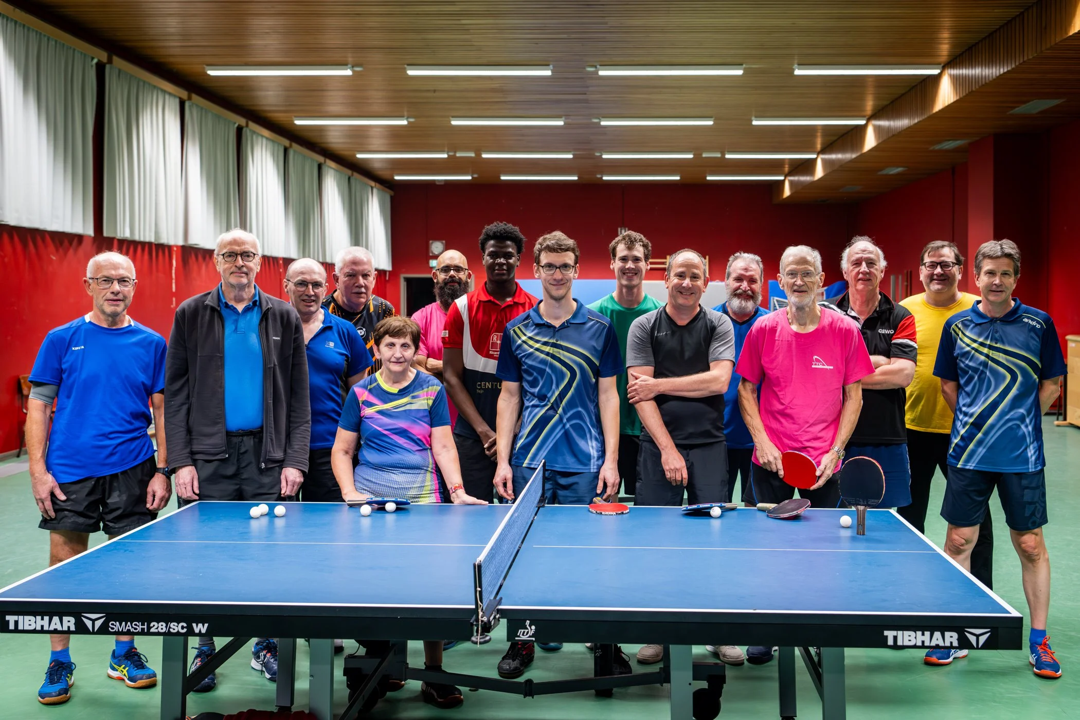 Man in glazen, donker jack en blauwe polo die een positieve houding toont met een duim omhoog en een gebalde vuist tegen een kleurrijke, bokeh achtergrond.