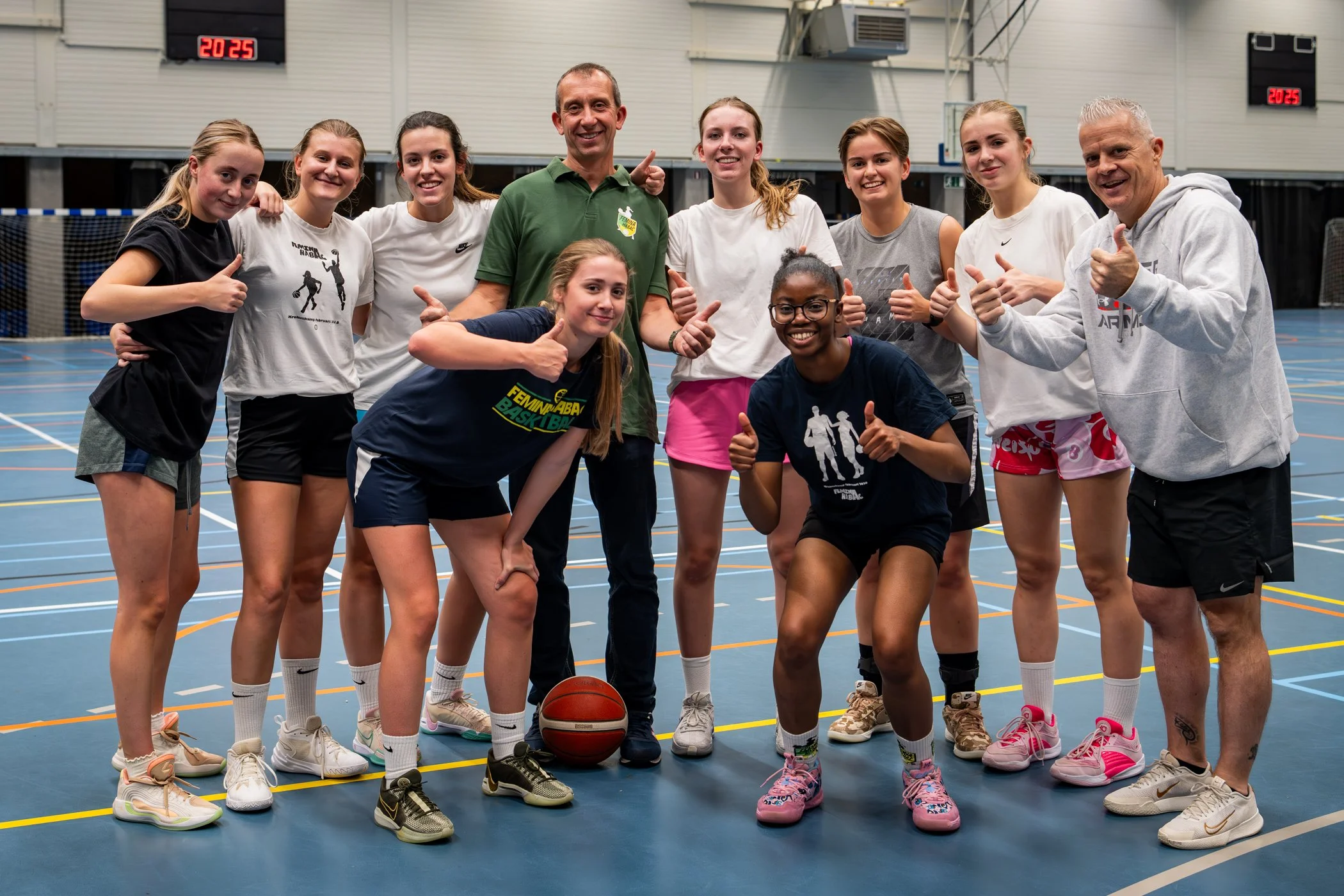 Vijftien mensen in groene en gele sportkleding en sjaals, glimlachend en handen gebaren zoals duimen omhoog, tegen een roze en paarse bokeh achtergrond.