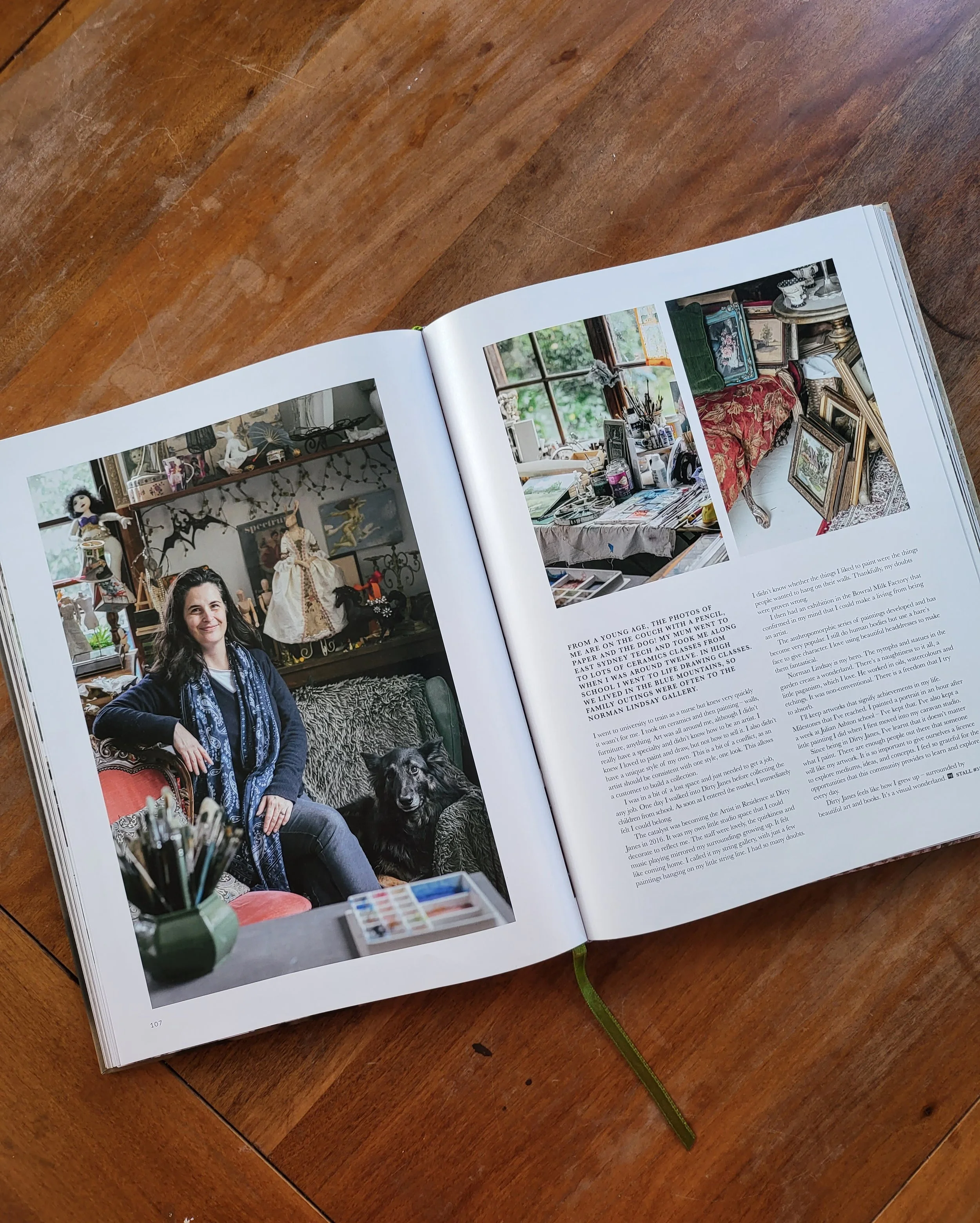 Open book displaying a photo of a woman sitting on a couch with a dog and art supplies, alongside images of an artist's workspace and framed pictures, on a wooden table.