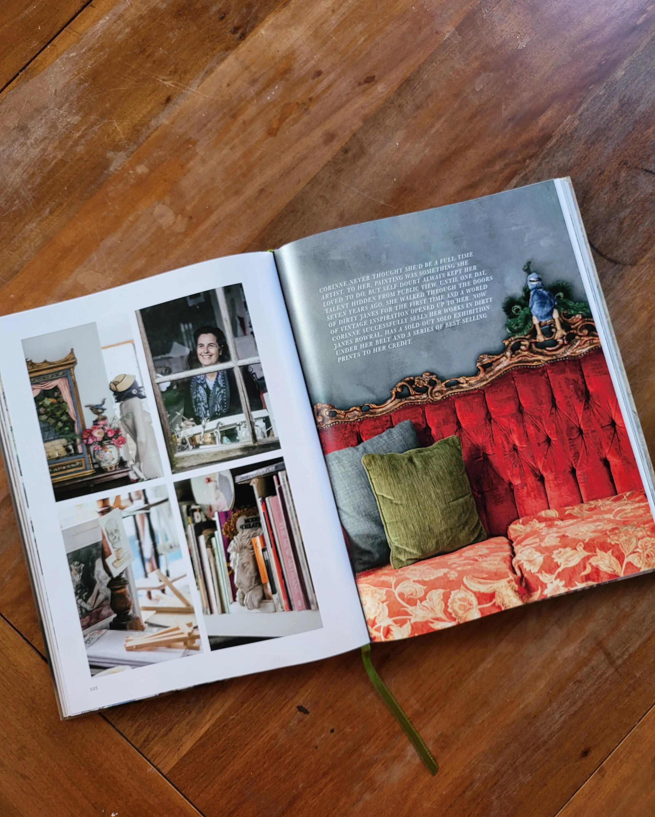An open book on a wooden table showing interior design. The left page has multiple images of a workspace with art supplies and decor. The right page features a text passage and a photo of a vintage red velvet couch with decorative pillows.