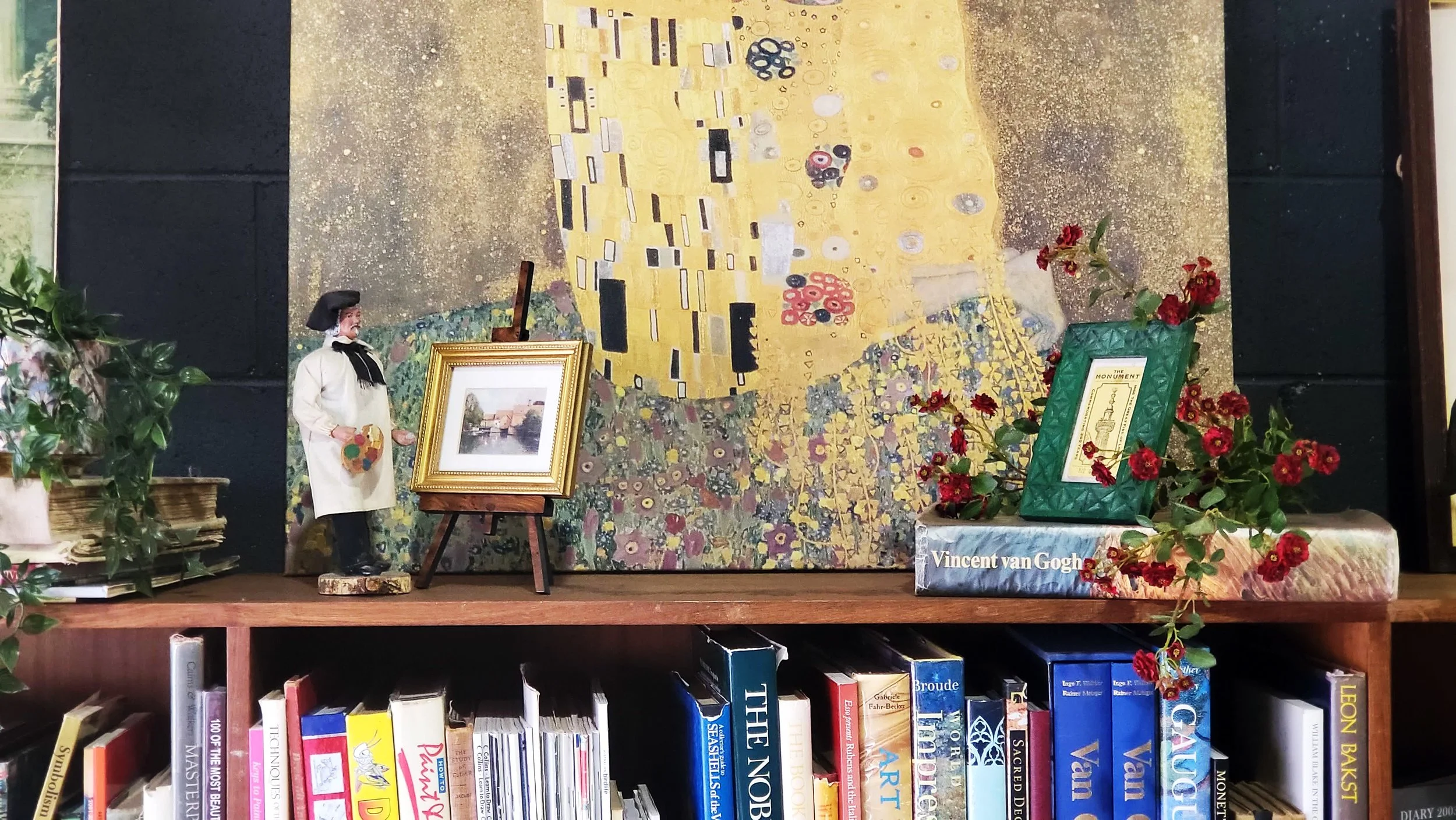 Bookshelf with art books, decorative painting, artist figurine, framed picture, and artificial red flowers.