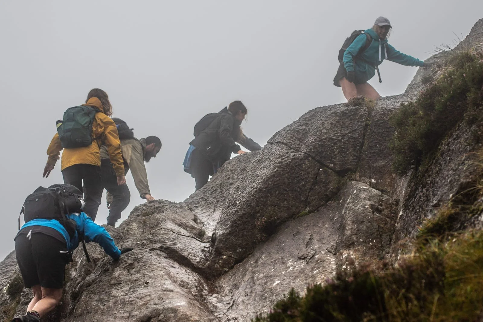 Group of hikers climbing a steep, rocky mountain in foggy weather, equipped with backpacks and outdoor gear.