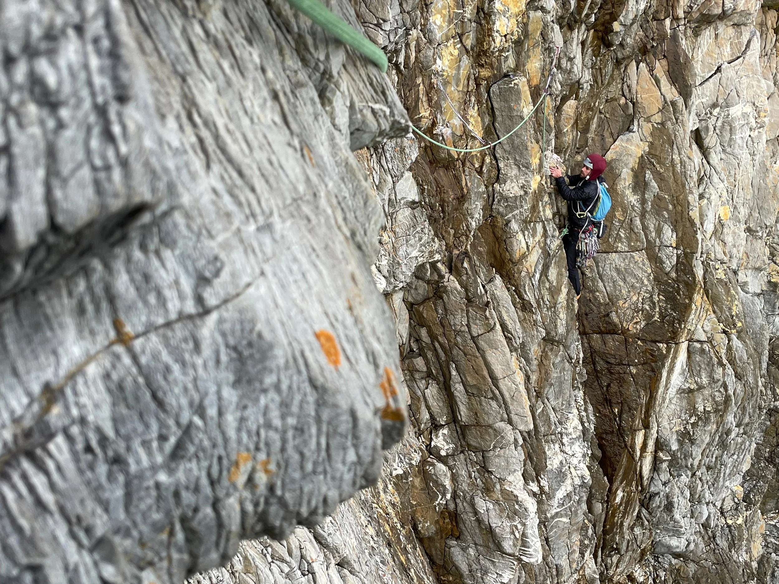 A person rock climbing on a rugged, steep cliff face, wearing a red helmet, black clothing, and carrying climbing gear, with a safety harness and rope attached.