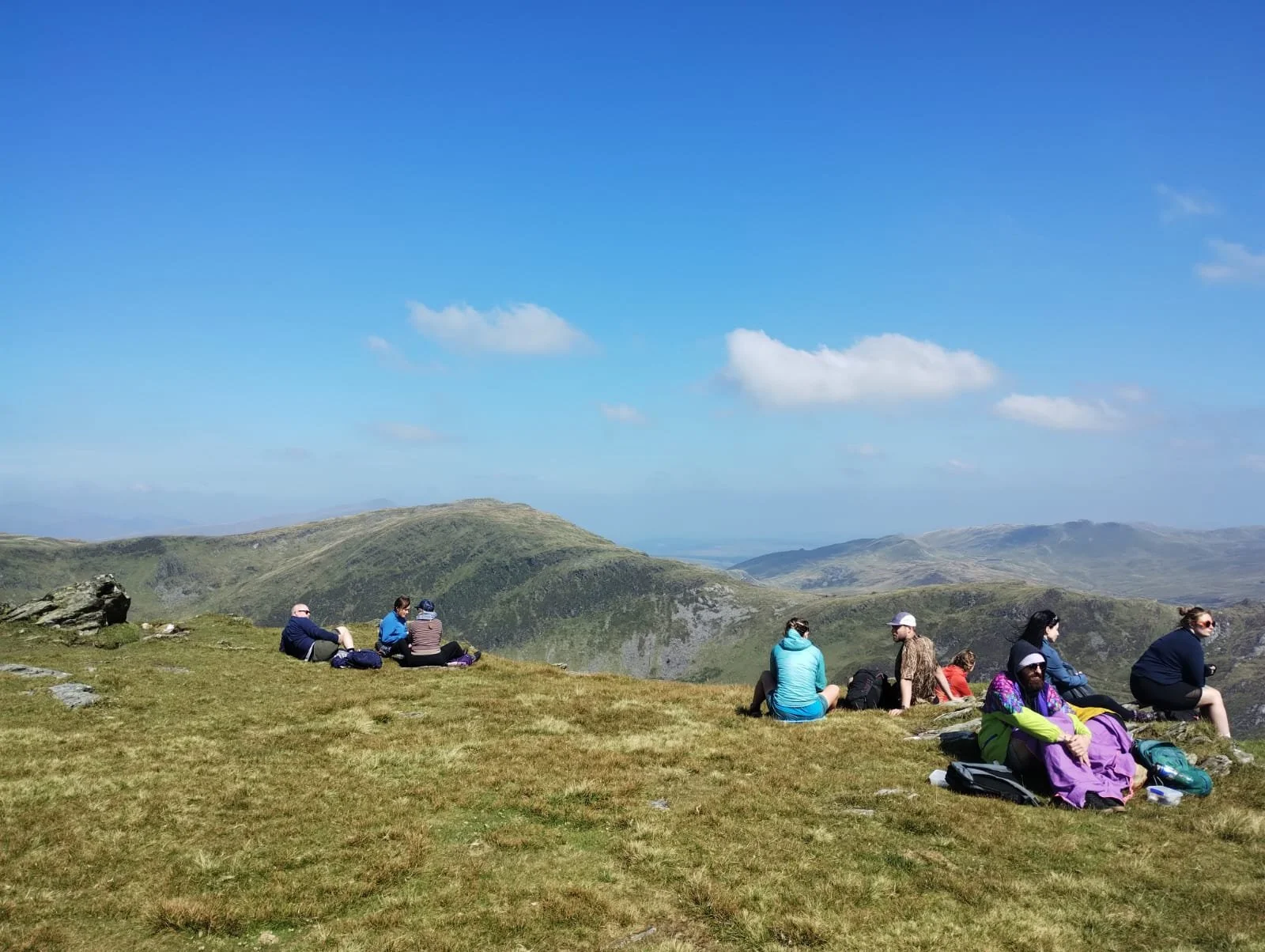 Group of people sitting on grassy hillside overlooking mountainous landscape and blue sky with wispy clouds.