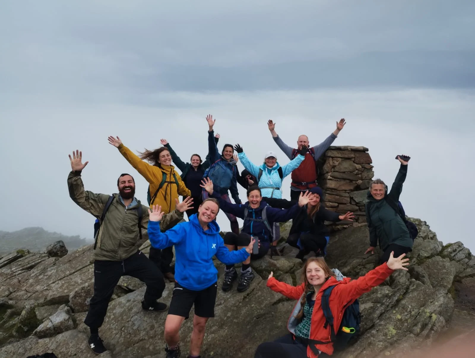 Group of ten hikers celebrating on a mountain summit with cloudy sky, some raising their arms and smiling.