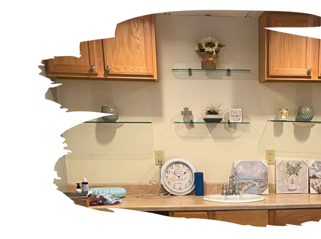 Kitchen countertop with a clock, a soap dispenser, and a blue tumbler, with wall shelves holding decorative items and cabinets above.