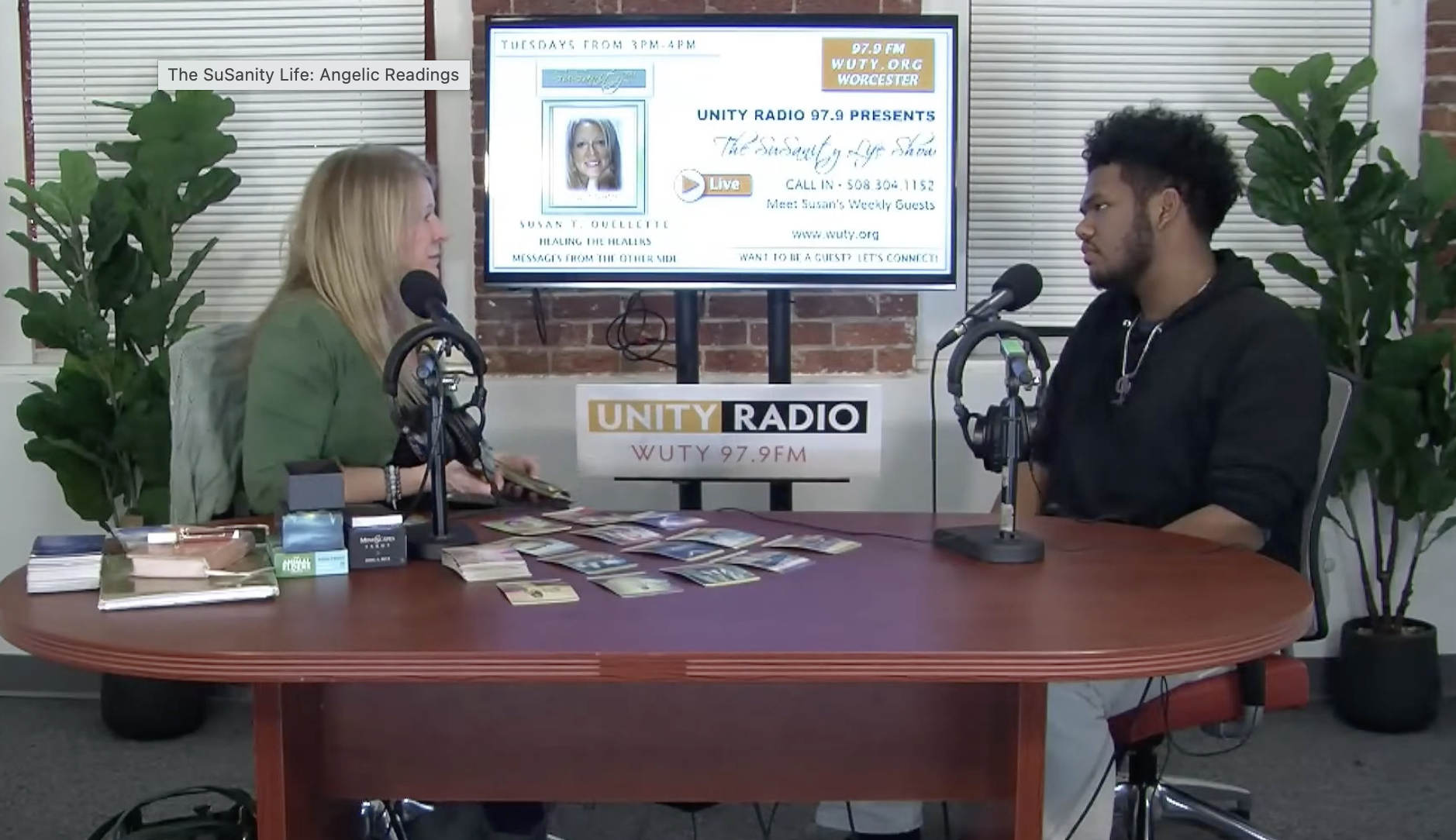 Two people sitting at a table in a radio studio. The woman on the left has blonde hair, and the man on the right has dark curly hair. The table has various tarot cards, books, and small items. Behind them is a screen displaying a radio show schedule, station information, and a photo of the woman. The studio has brick walls, potted plants, and a UNITY RADIO sign on the table.