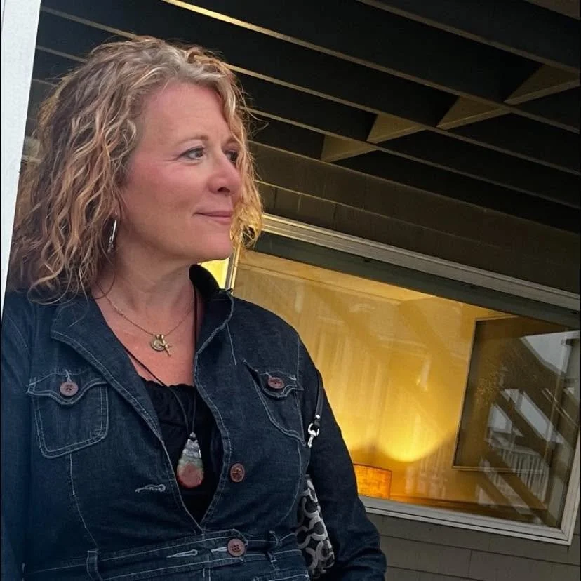 A woman with curly blonde hair looking to her left, standing outdoors near a building with a wooden wall and large window.