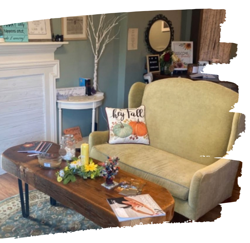 Living room with a light green armchair, a wooden coffee table with holiday decorations, and a decorative pillow with fall pumpkins and the text "Hey Fall." In the background, there is a white fireplace mantle, framed pictures, a small white side table with a white decorative tree, and various framed artwork and decor on the wall and shelf.