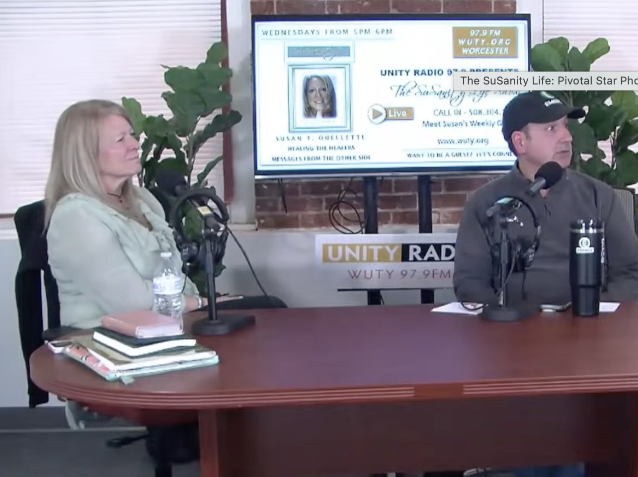 A woman and a man are sitting at a table during a radio broadcast. The woman is on the left, wearing a light-colored jacket, with a water bottle, notebook, and stacks of papers in front of her. The man is on the right, wearing a dark jacket and baseball cap, with a water bottle and microphone in front of him. Behind them is a large screen displaying information about the show, with green plants on each side.