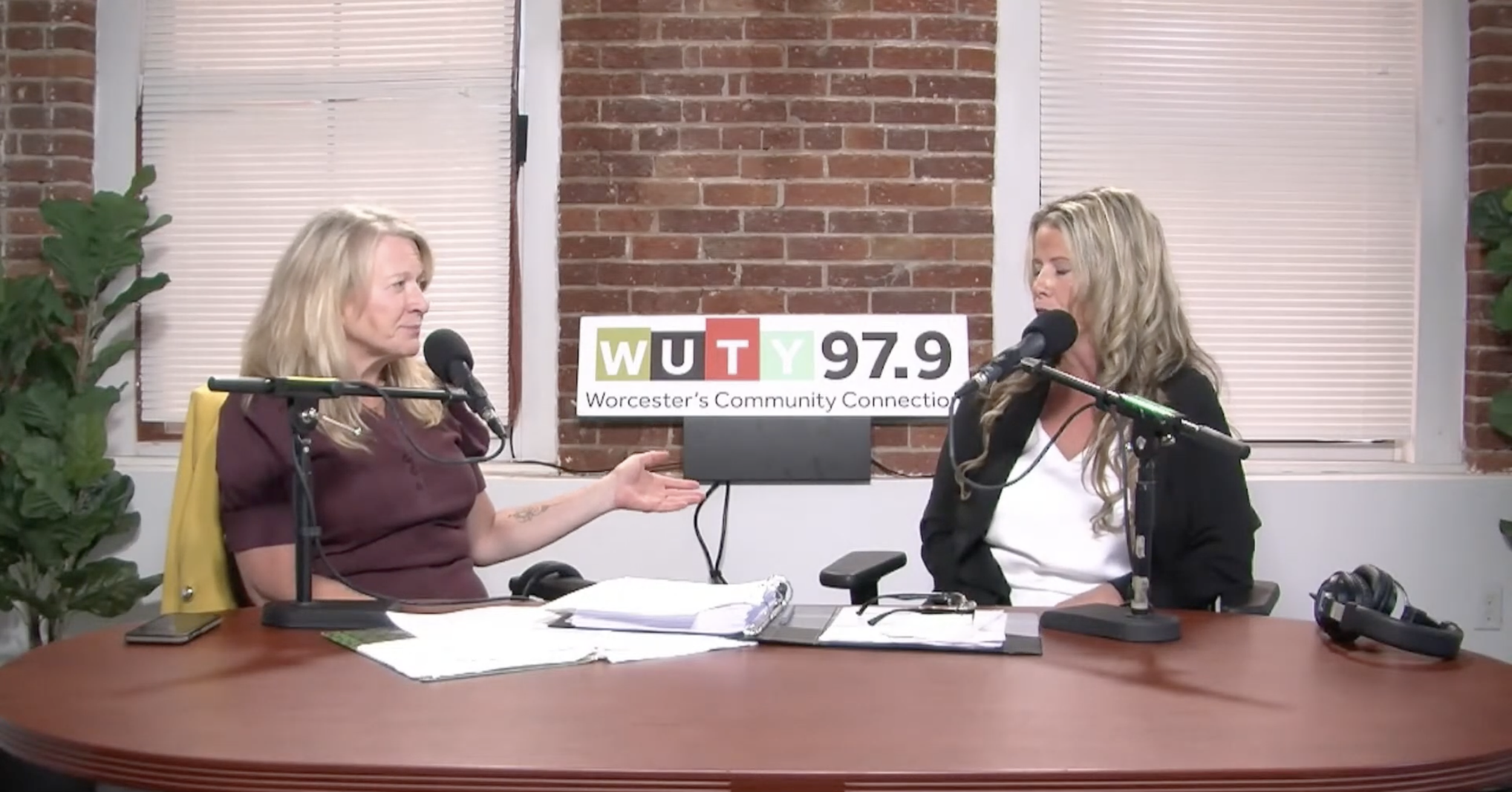 Two women are seated at a table in a radio station studio, engaging in a conversation. One woman is gesturing with her hand, while the other is listening. There are microphones, papers, and electronic devices on the table. Behind them is a brick wall with windows and a sign that reads "WUTY 97.9 Worcester's Community Connection."