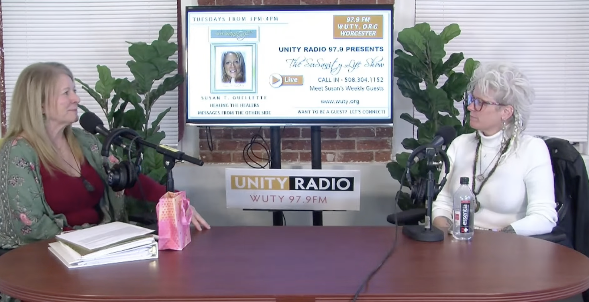 Two women are seated at a table in a radio studio, engaged in conversation. Behind them, a large screen displays information about a radio show called 'The SusSusan Age Show' on Unity Radio 97.9 FM, with details for calling in and visiting the website. The studio has large green plants and a brick wall. One woman has long blonde hair, is wearing a green floral jacket over a red top, and has documents in front of her. The other woman has short, curly, gray hair, wears glasses, a white turtleneck, and has a water bottle and microphone in front of her.