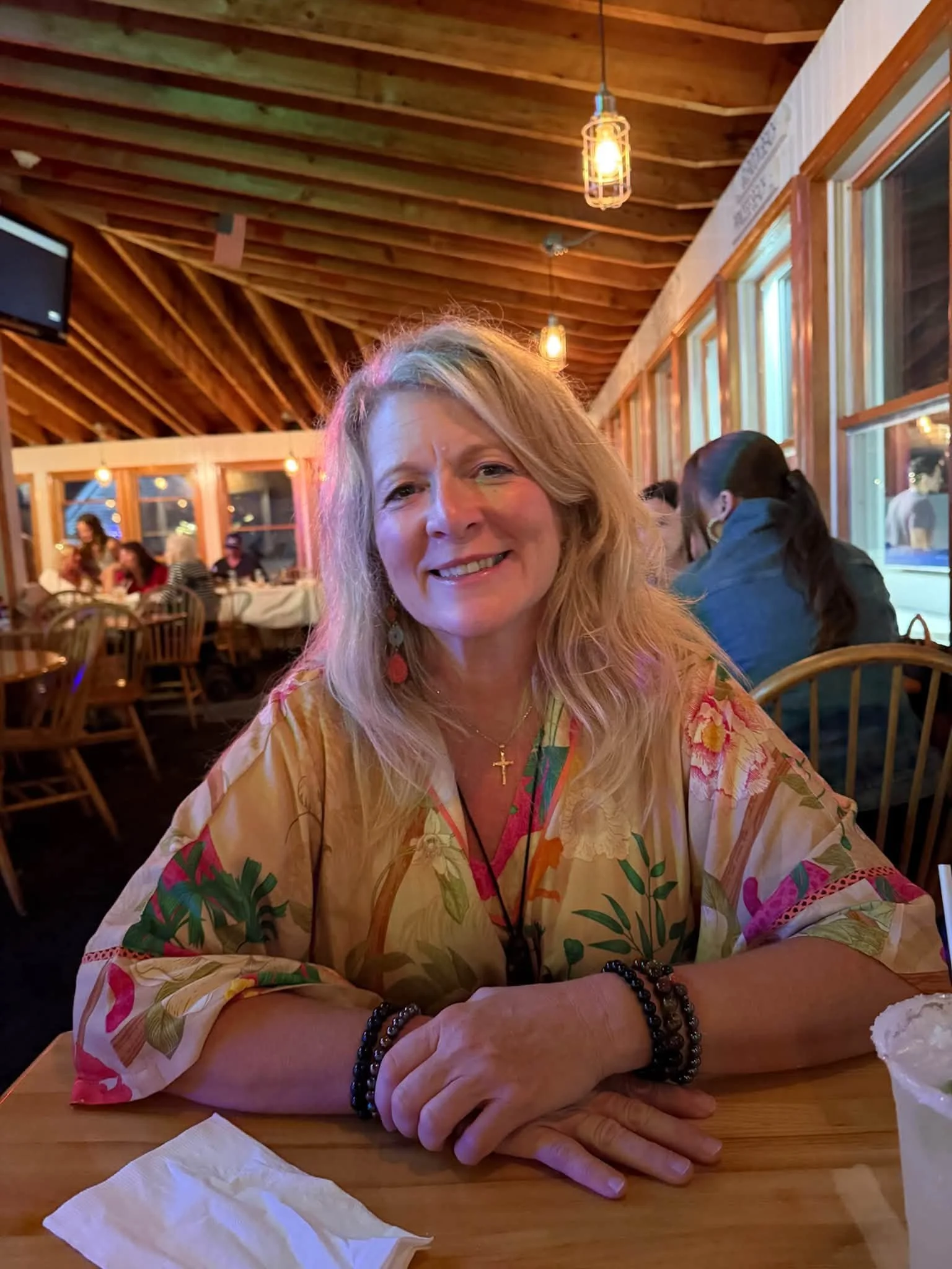 A smiling woman with long blonde hair sitting at a wooden table in a restaurant with a rustic ceiling. She is wearing a colorful, floral-patterned blouse, gold cross necklace, and black beaded bracelets on her right wrist. In the background, there are other people dining and large windows with wooden trim.