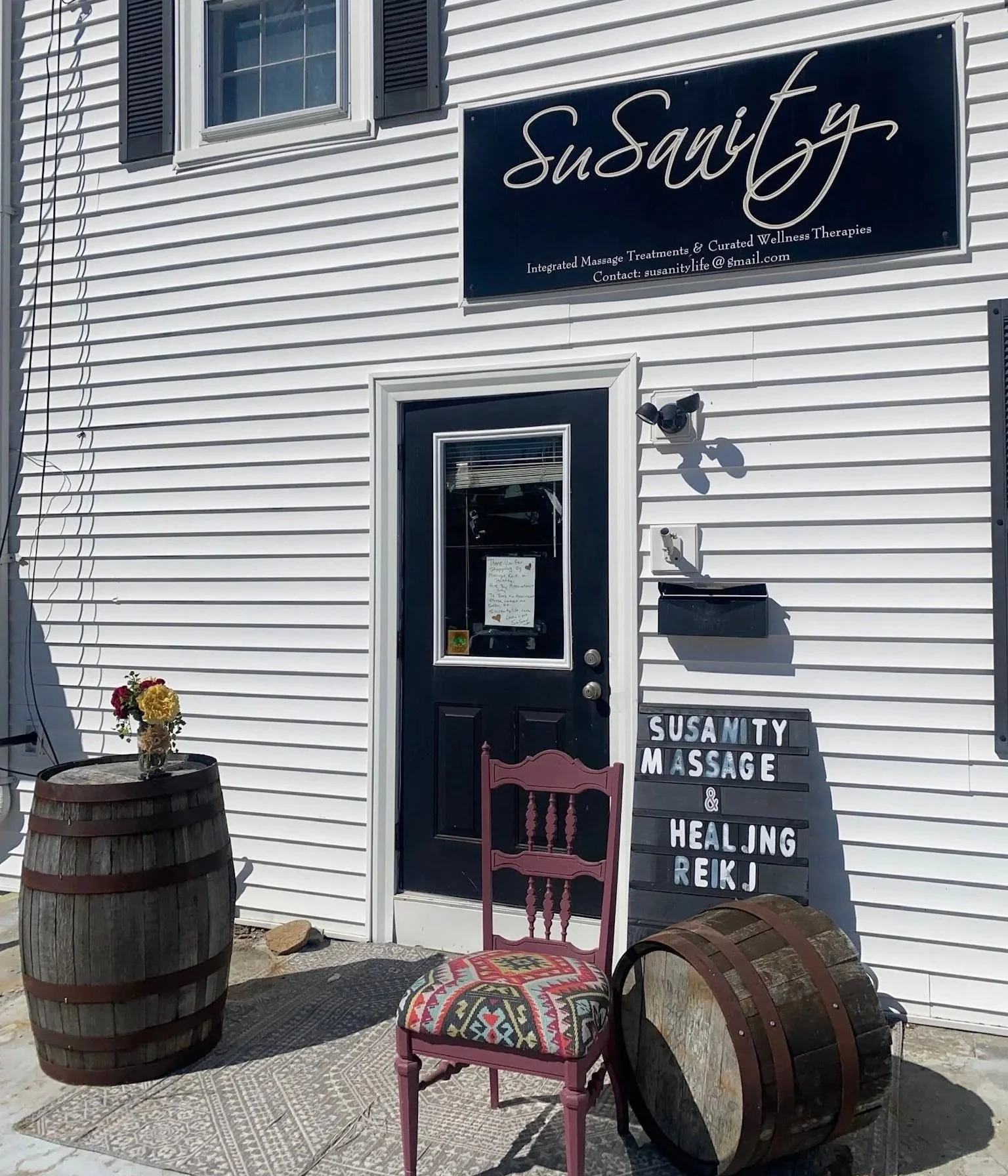 Exterior of a wellness therapy business with a sign reading "Susanity" and contact email. Decor includes flower arrangements in barrels, vintage and colorful chairs, and a sign indicating massage, healing, and Reiki services.