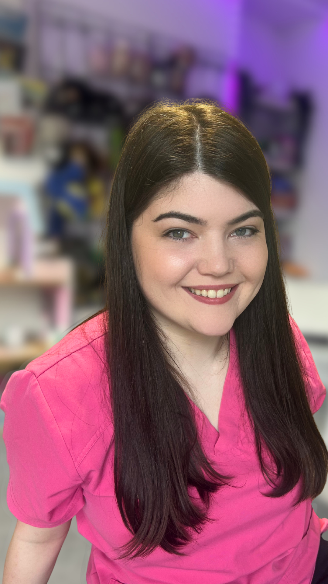 A woman with long dark hair and green eyes wearing a pink medical scrub top, smiling in a colorful workspace.
