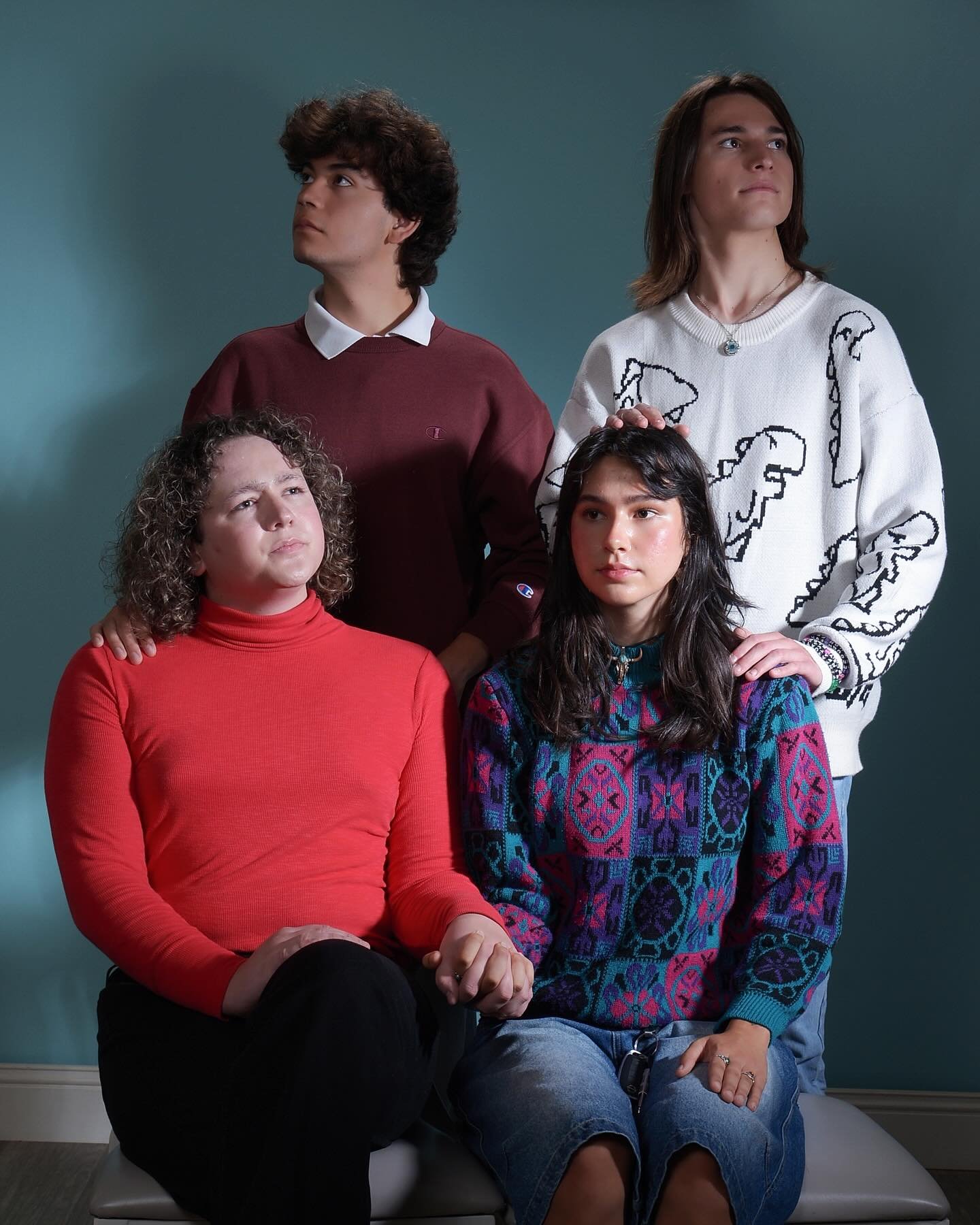 Four people, two men and two women, pose against a teal background. One woman is seated with her hands clasped, dressed in a red turtleneck, and another woman is seated with her hands on her lap, dressed in a colorful patterned sweater and ripped jeans. Behind them, a man in a white shirt and burgundy sweater, and another man in a white sweater with black patterns, stand with their hands on the seated women’s shoulders.