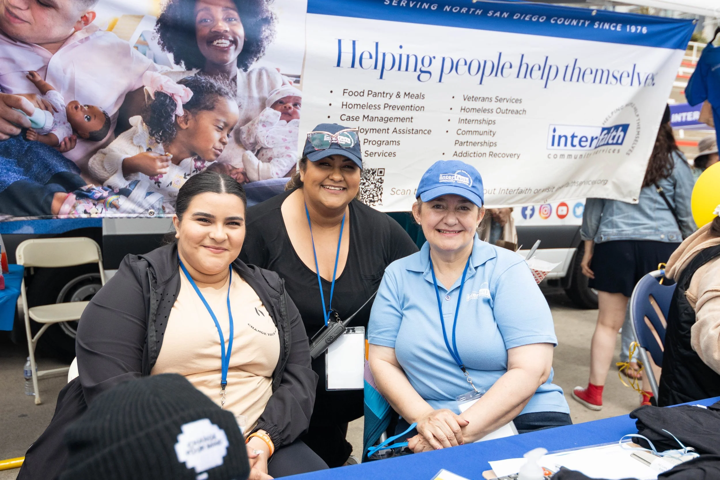 Three women sitting at a table in front of a banner for Interfaith community services, smiling for the camera. The banner behind them features images of diverse people and a list of services offered.