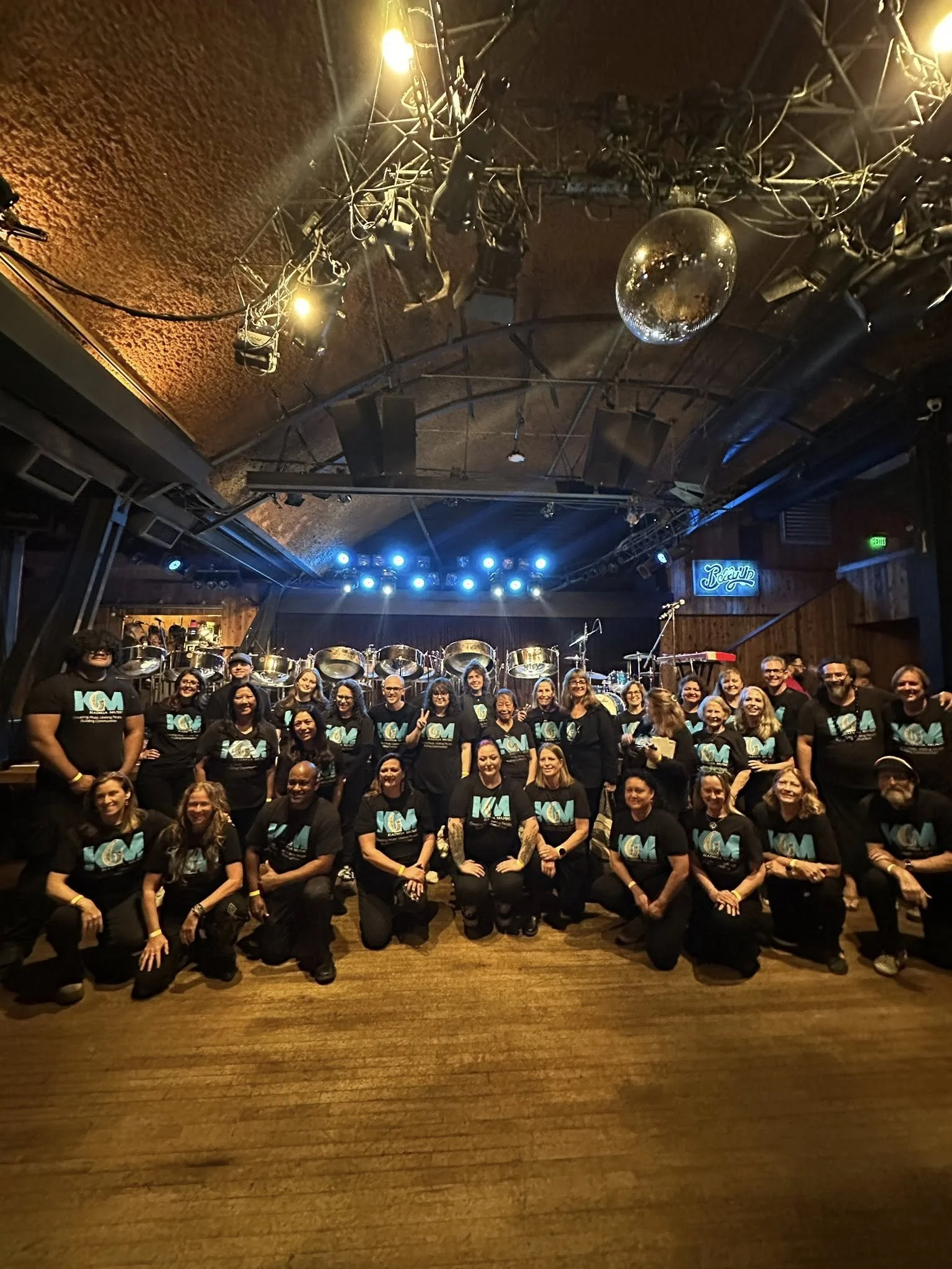 Group of people, including men and women, posing together on a stage or dance floor with musical instruments and blue stage lights in the background. The setting appears to be a concert or performance venue.