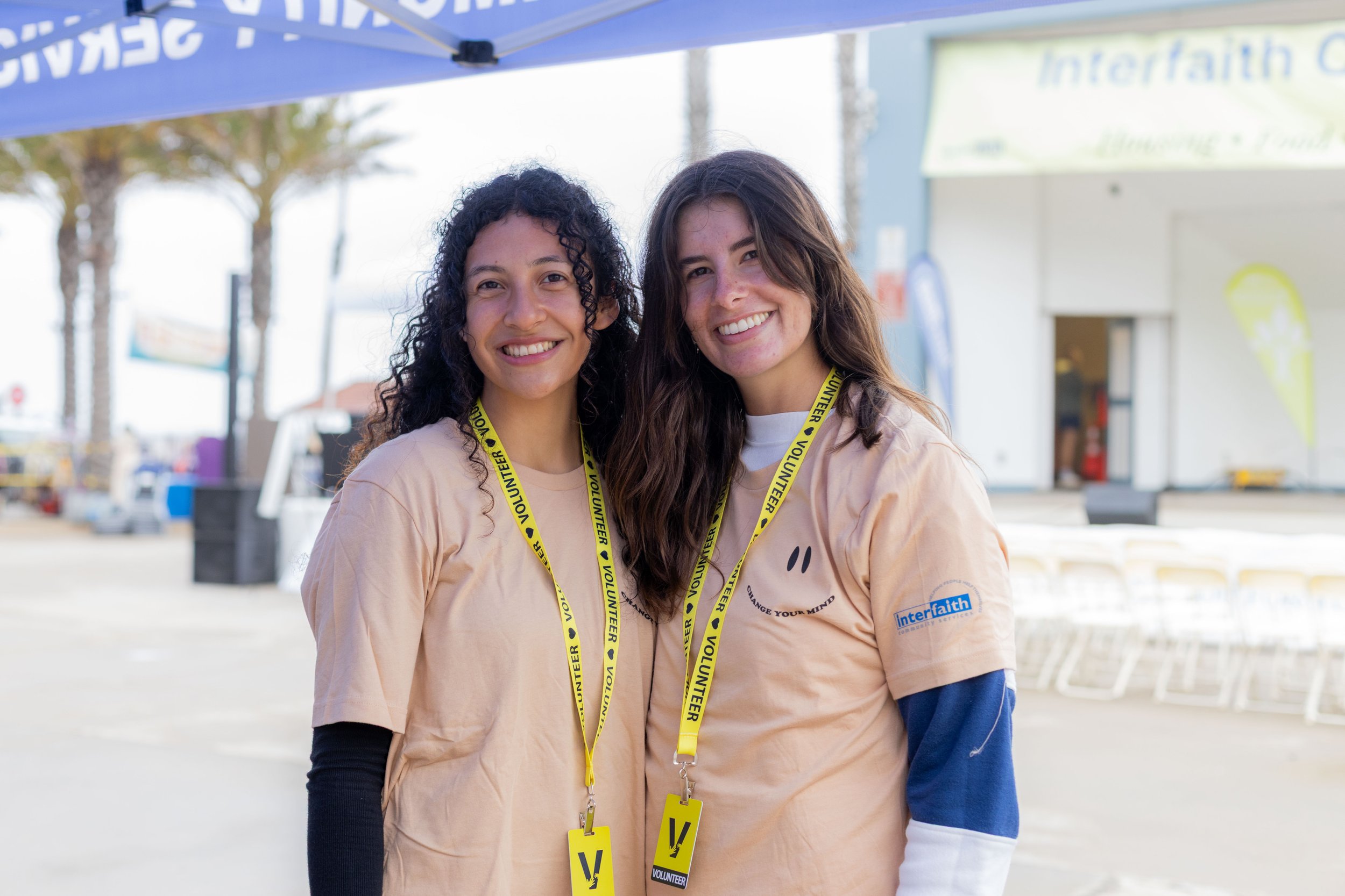 Two young women smiling at the camera, wearing beige volunteer T-shirts and yellow volunteer lanyards, standing outdoors in front of a tent or booth with a blue canopy.