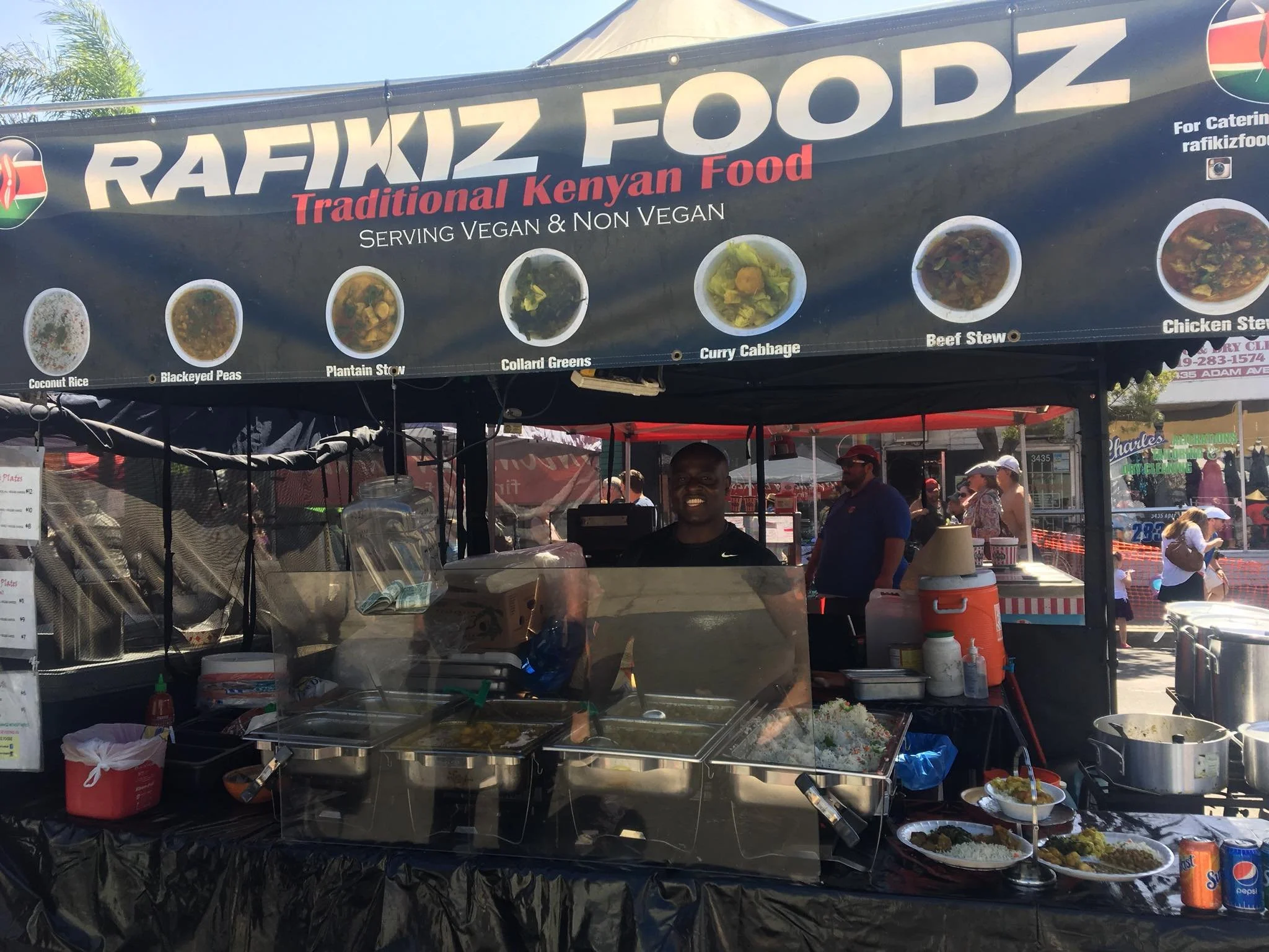 Food stand with a sign that reads 'RAFIKZ FOODZ Traditional Kenyan Food Serving Vegan & Non Vegan' and pictures of dishes like coconut rice, black-eyed peas, plantain stew, collard greens, curry cabbage, beef stew, and chicken stew, with people working behind the counter at an outdoor event.