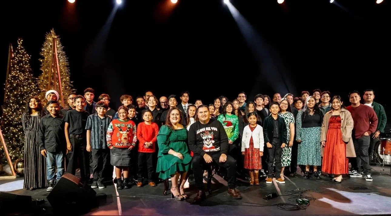 Group of children and adults on stage for a holiday celebration, with Christmas trees decorated with lights in the background.