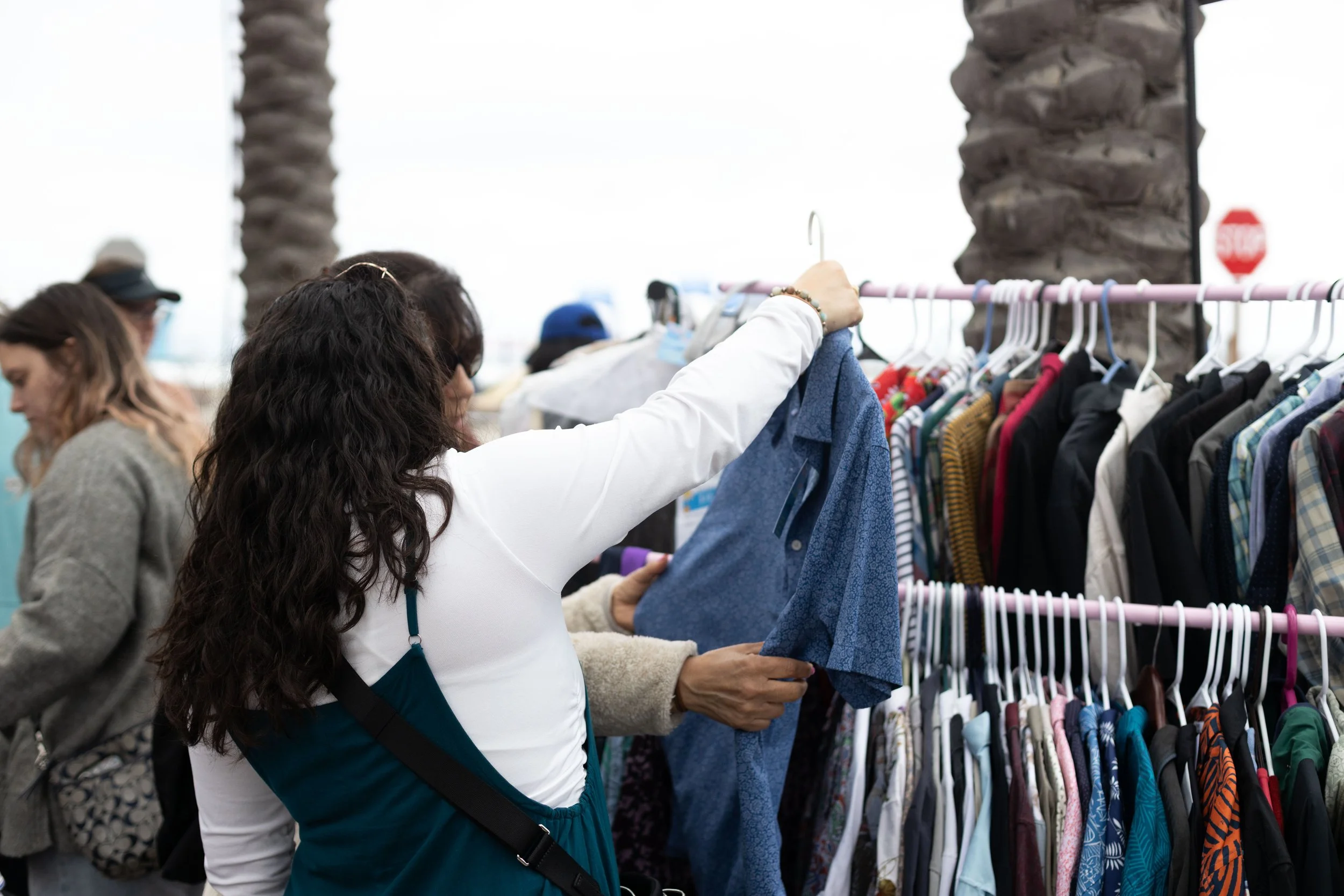 People shopping for clothes at an outdoor market with racks of clothing and palm trees in the background.