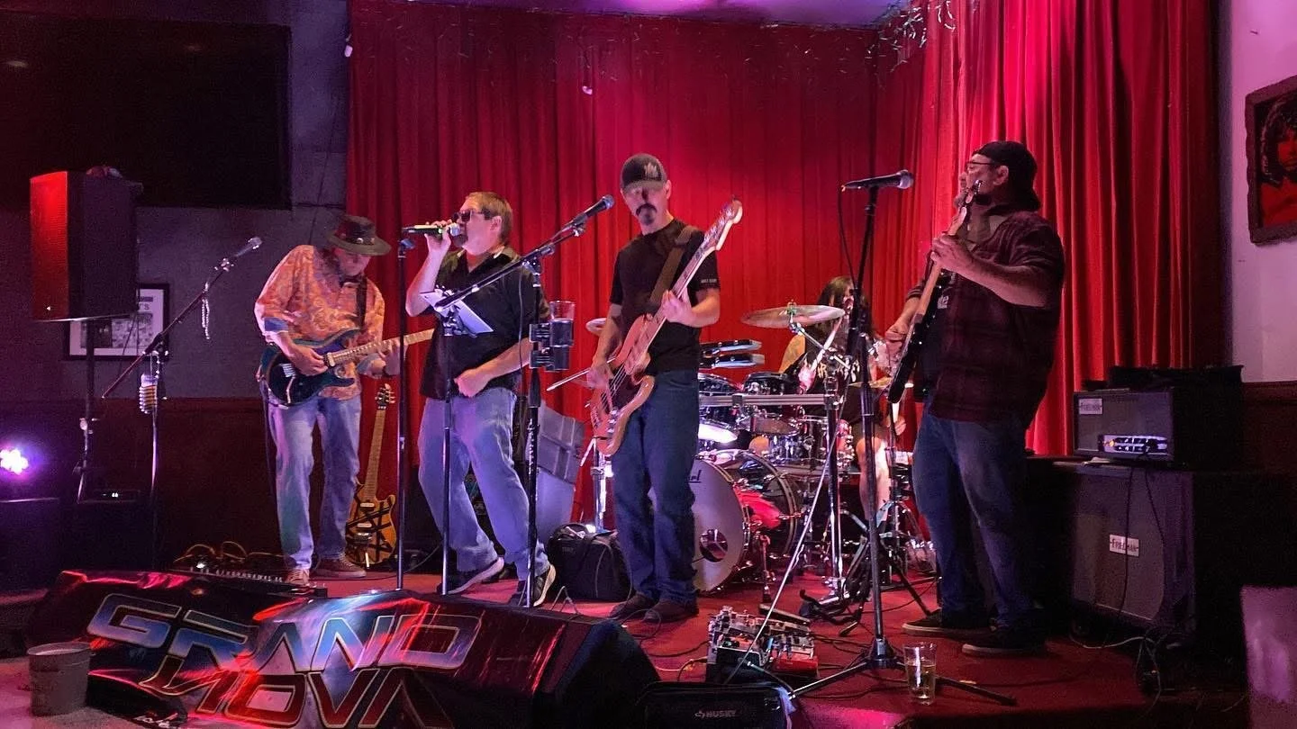 A band performing on stage with red curtains in the background. The band has five members: a guitarist wearing a hat, a singer with a microphone, a bassist, a drummer, and a keyboard player. There are various musical equipment and speakers on stage.