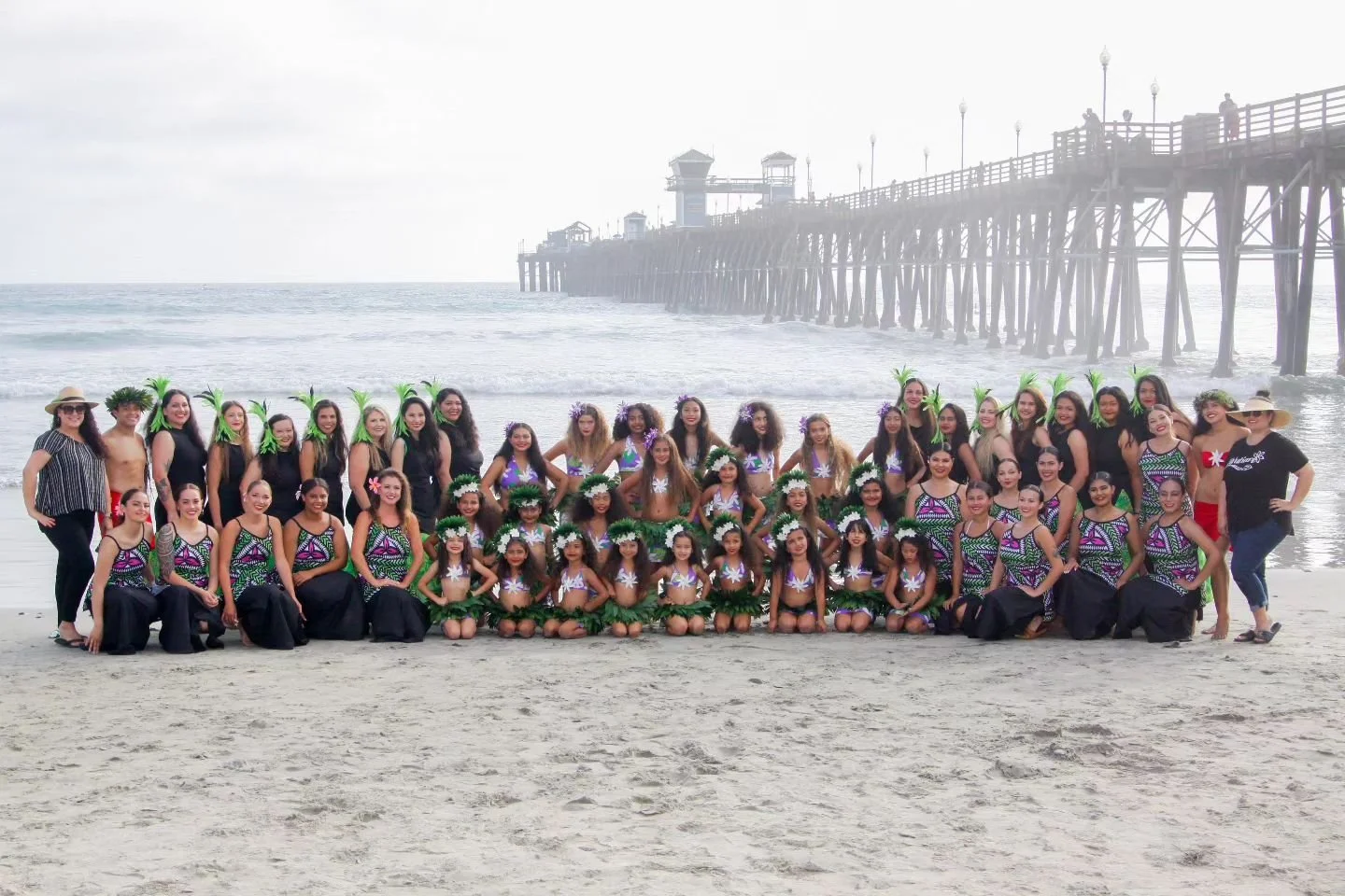 Group of women and children dressed in Hawaiian-themed costumes, standing on a beach with a pier in the background.