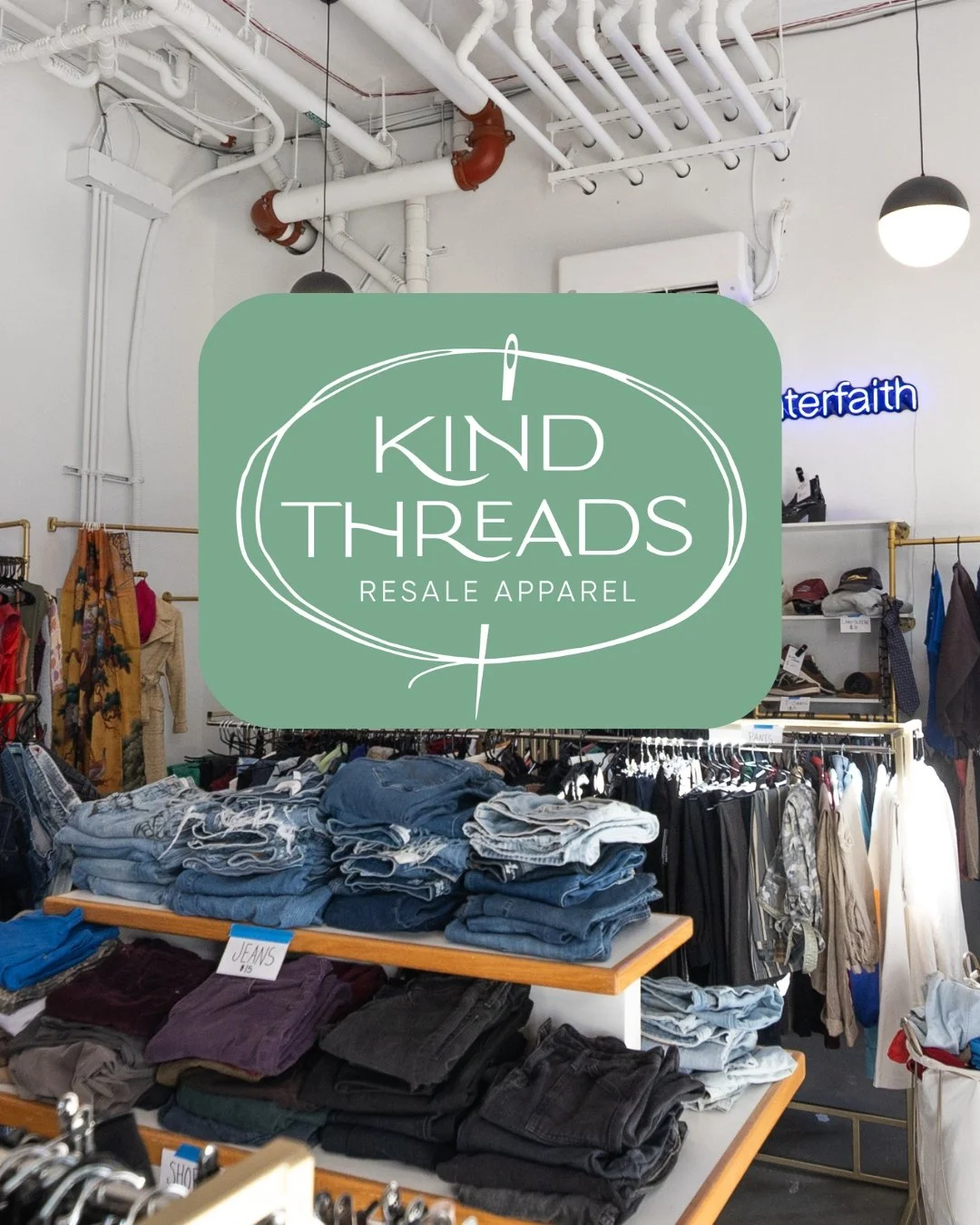 Inside a thrift clothing store with folded jeans on a table, racks of clothing on hangers, and a neon sign in the background.