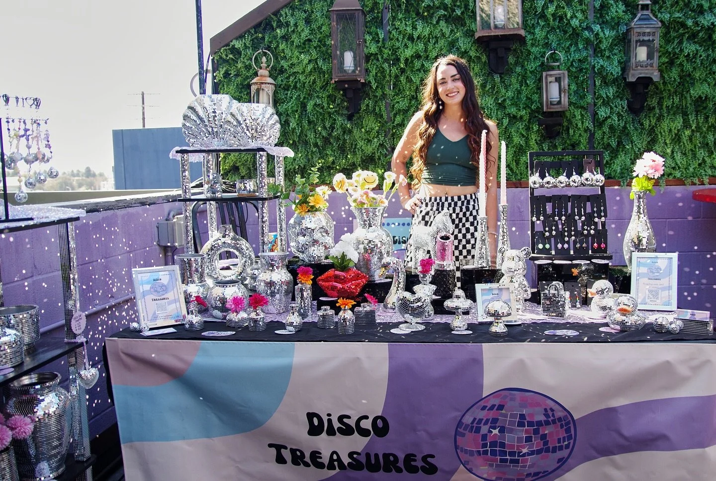 A young woman with long curly dark hair smiling at a jewelry display table at an outdoor event. The table has various shiny, reflective jewelry pieces including rings, necklaces, and earrings made with disco ball-like metallic designs. There are also flowers in vases and frames on the table, with a purple and green leafy background behind her.
