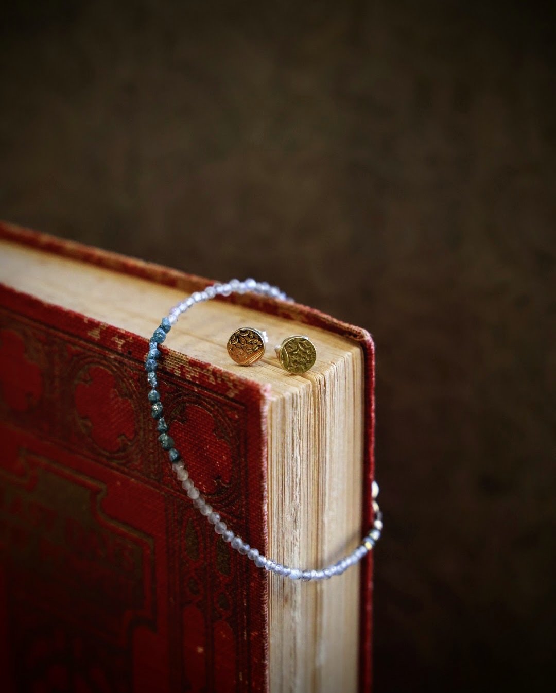 A beaded necklace and a pair of gold and silver earrings placed on top of a vintage red hardcover book with gold accents.